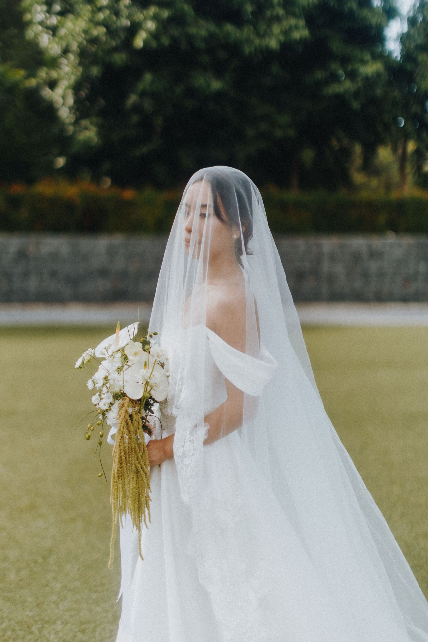 A bride in a flowing white dress and veil holds a white and gold bouquet on a grassy lawn.