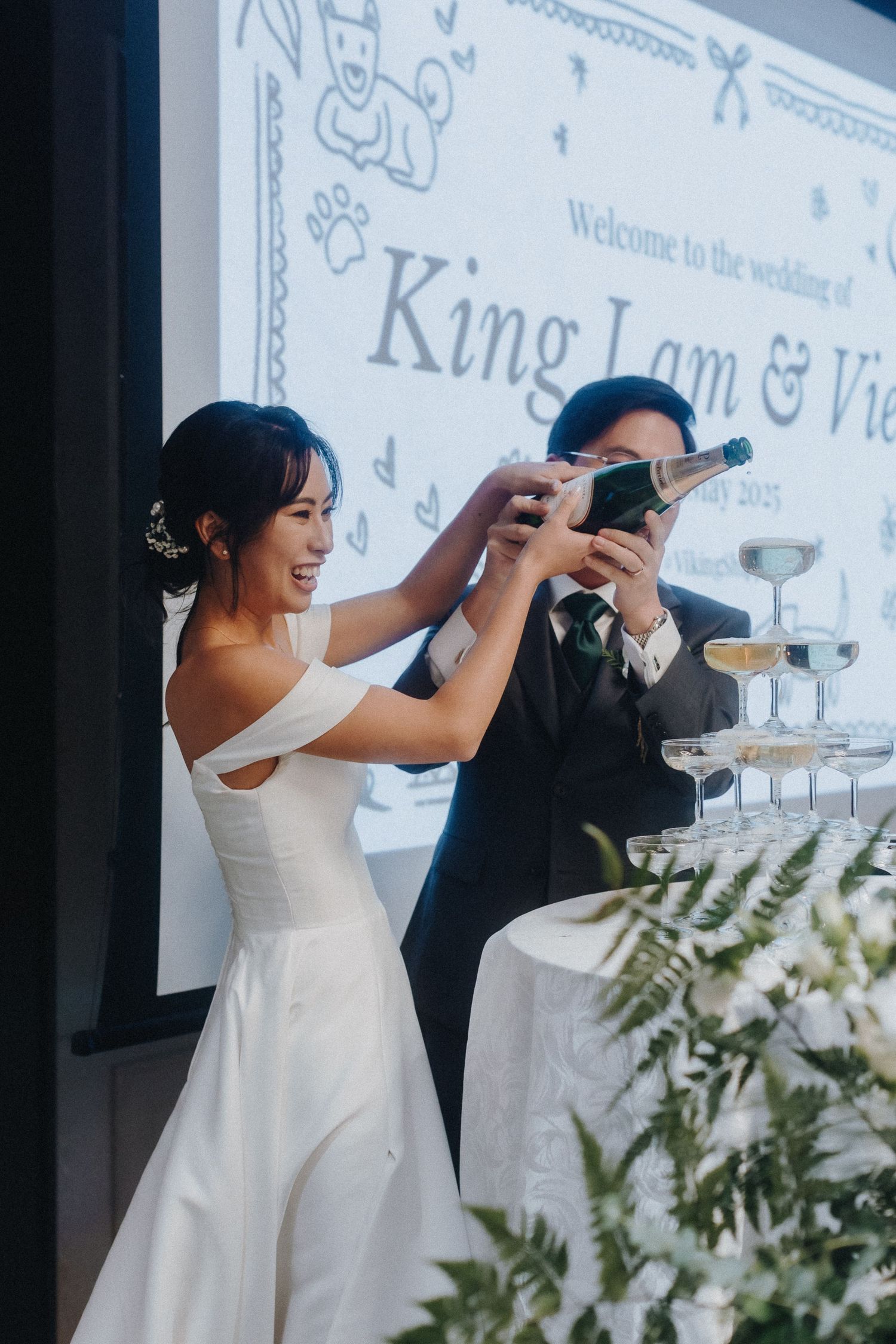 Couple pours champagne into tower of glasses at their wedding celebration.