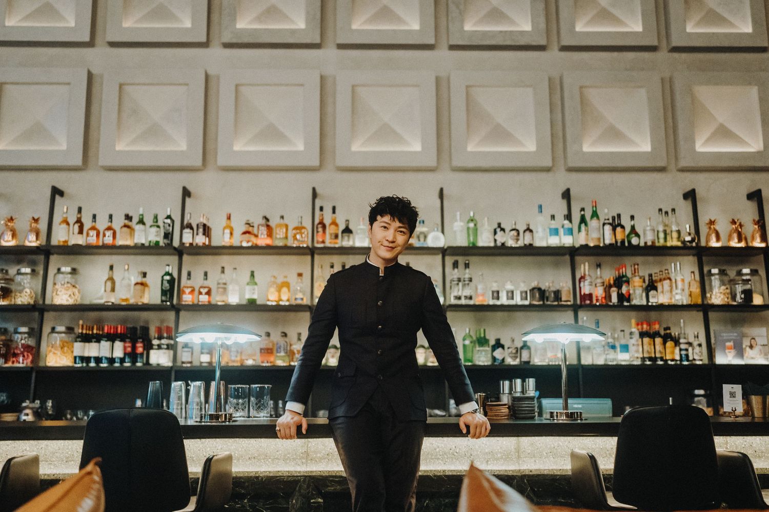 A bartender stands confidently in front of an impressive wall of bottles at an upscale bar.