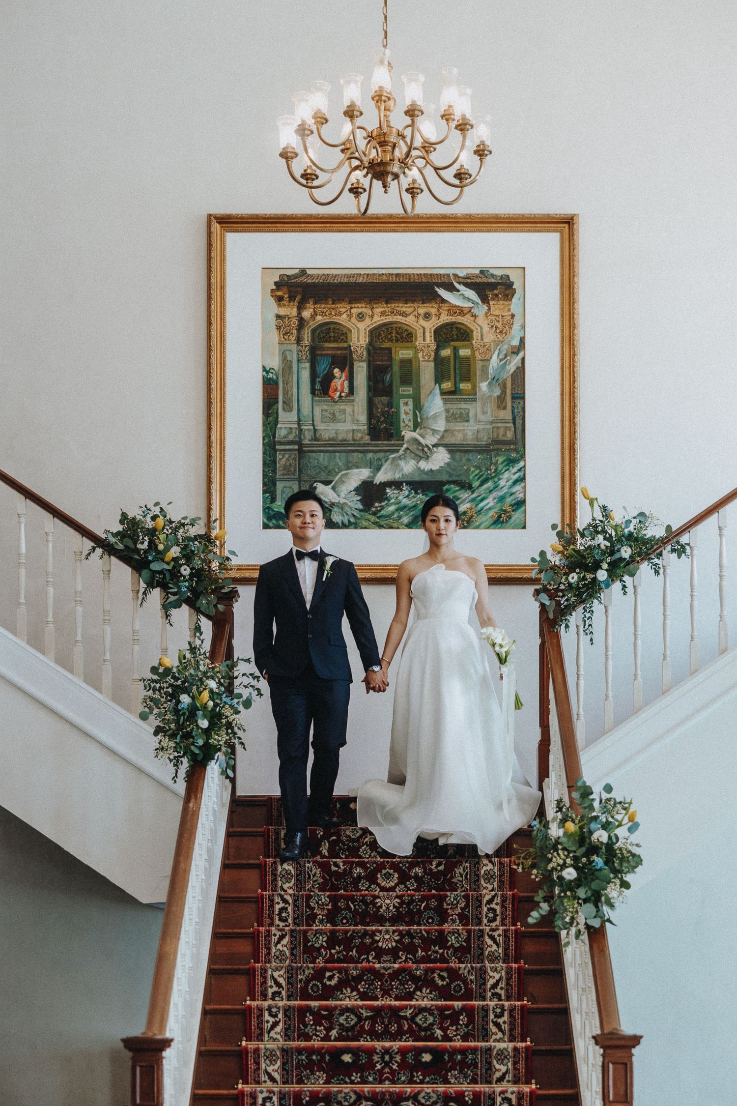 An elegant couple poses on a carpeted staircase decorated with floral arrangements beneath a grand chandelier.