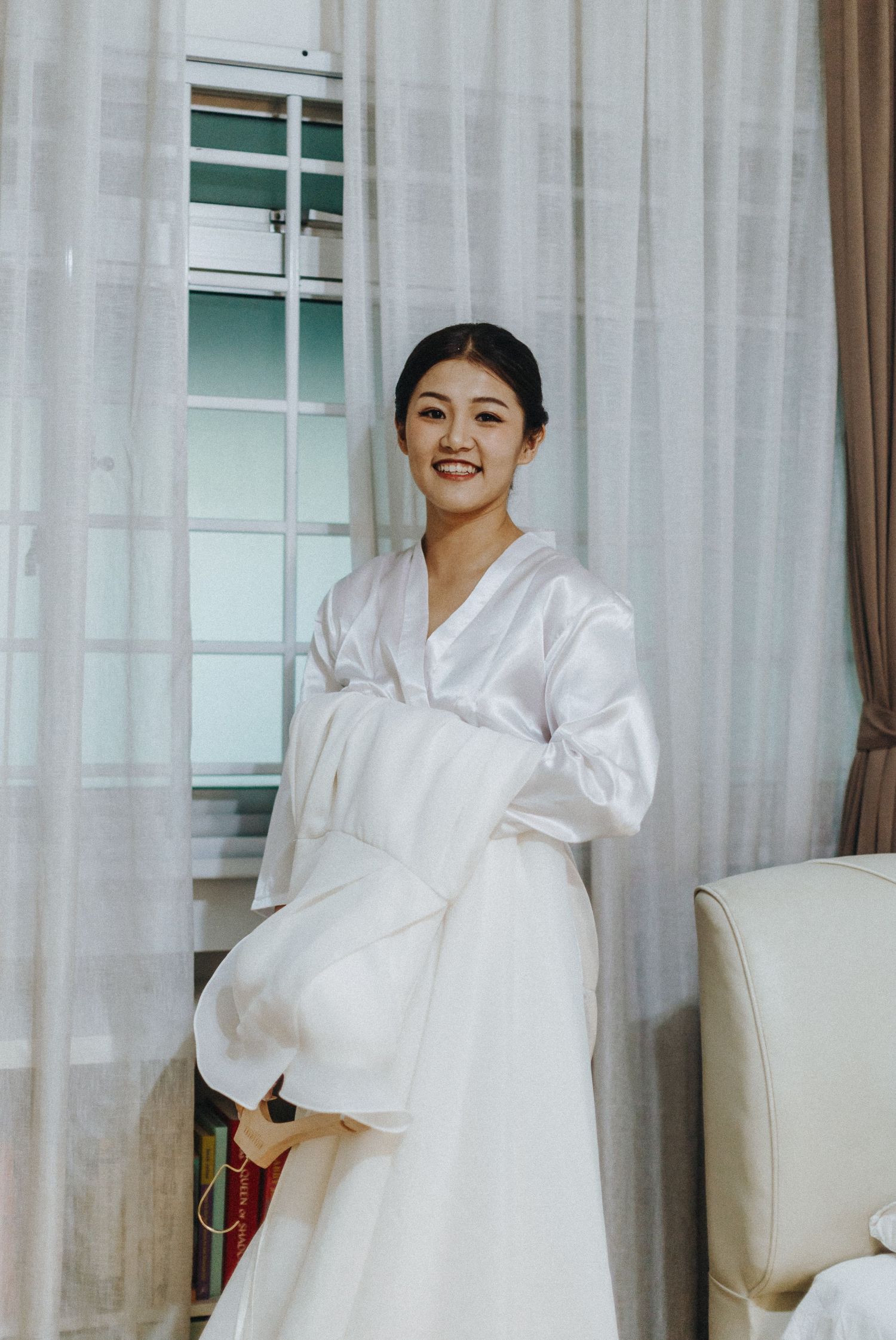 Bride getting ready in white robe before wedding ceremony with elegant curtains in background.