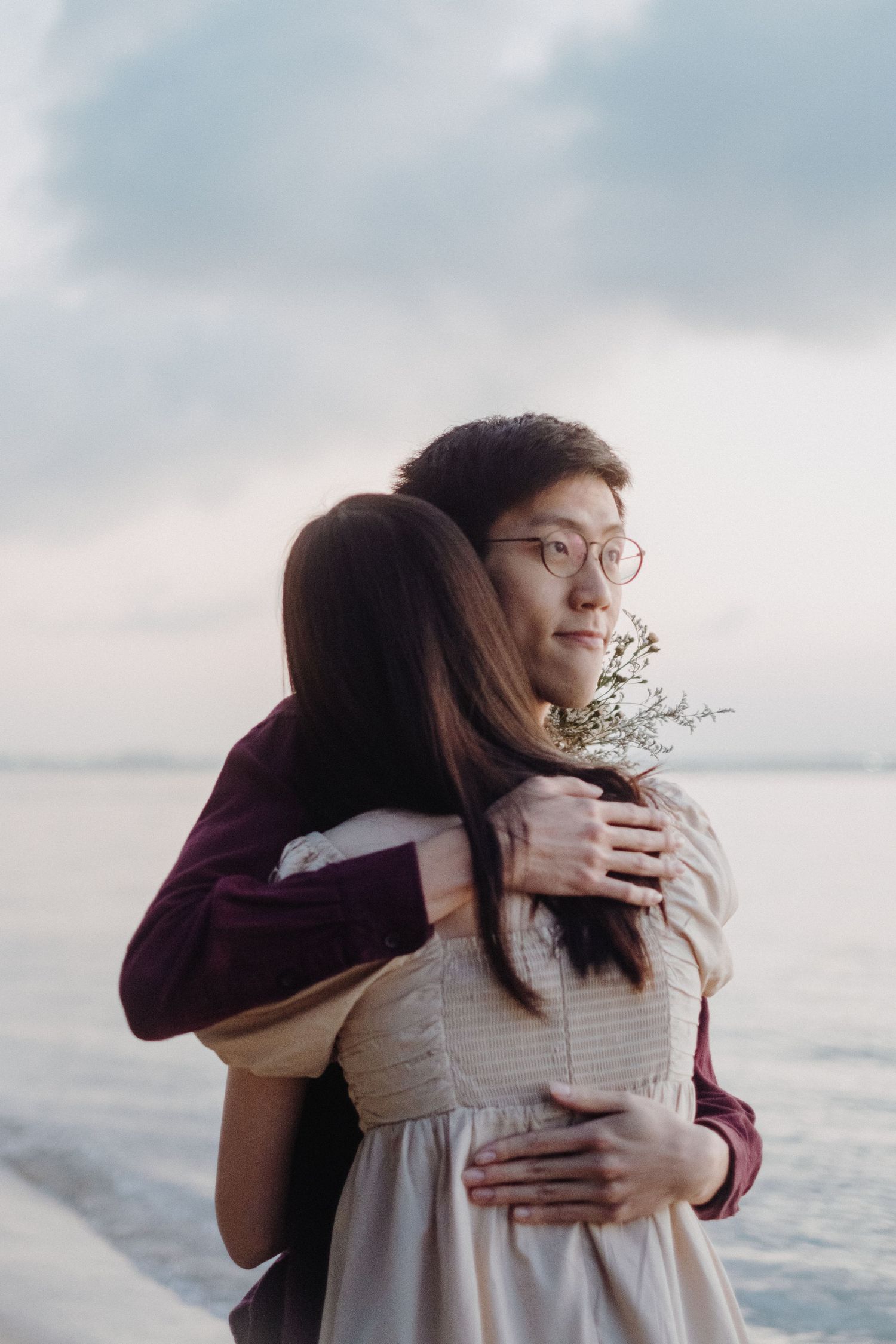 A tender moment captured at sunset showing a warm embrace by the water with soft cloudy skies in the background.