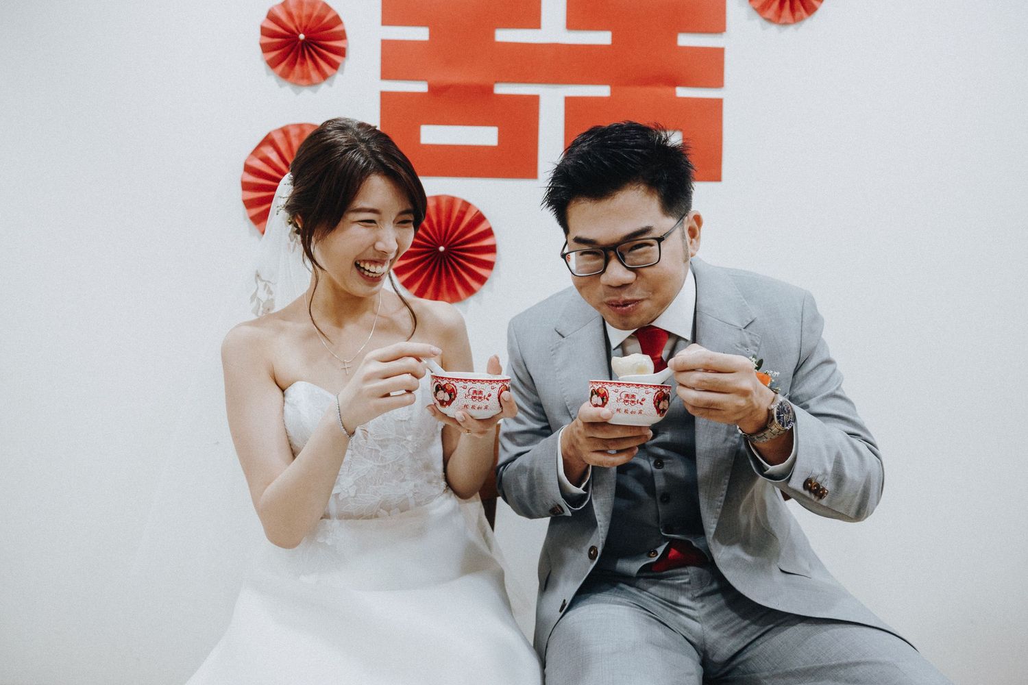 Newlywed couple in traditional wedding attire share tea together in front of red decorative fans on white wall.