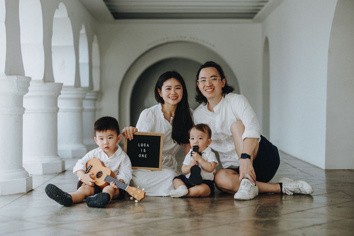 Family wearing white sits together on a light floor in an elegant arched colonnade hallway.