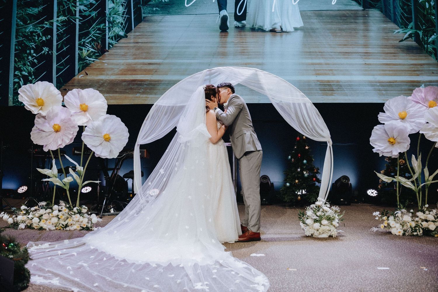 A wedding couple poses under a floral arch with large pink flowers and white drapery on a wooden deck.