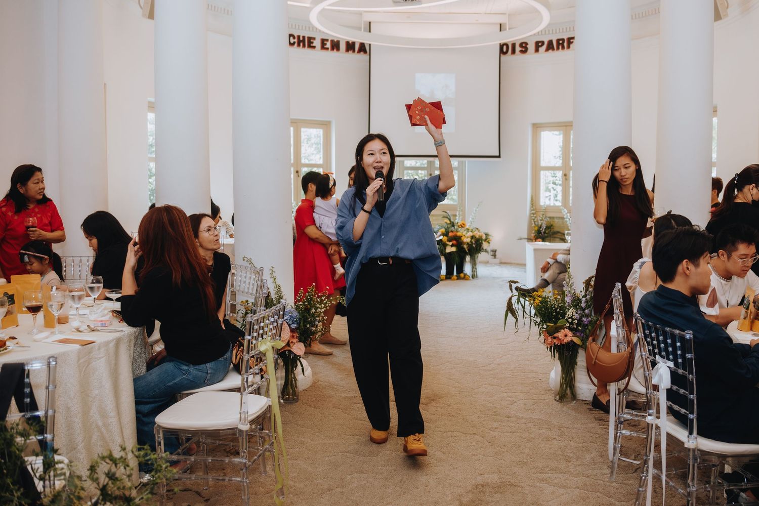 Wedding emcee speaks into microphone while guests dine at a decorated reception hall with white columns.