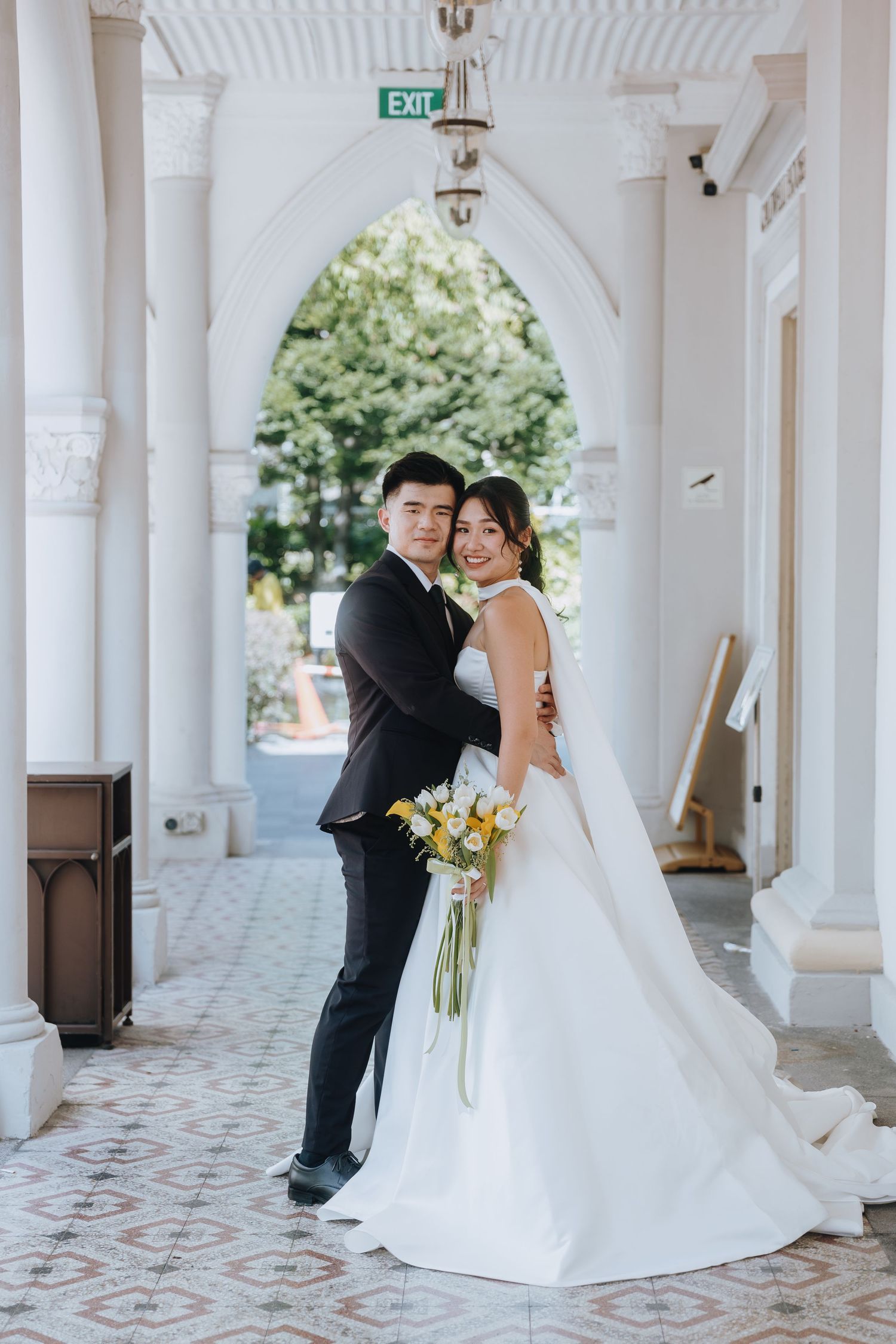 A smiling Asian couple in wedding attire pose under white arches, bride holding yellow bouquet in elegant white gown.