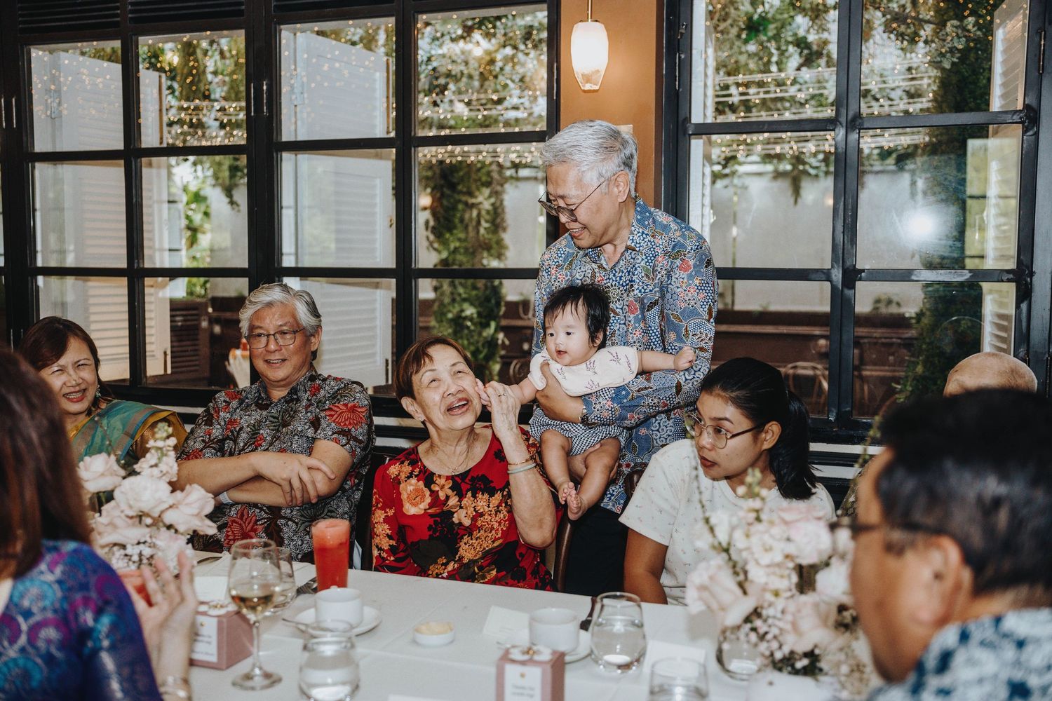 Elderly family members laugh joyfully with a baby at a warm and festive wedding reception.