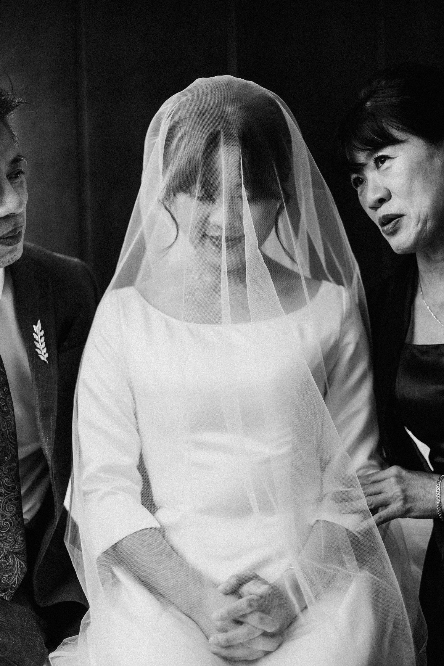 Black and white photo of a smiling bride in white dress and veil flanked by her parents on wedding day.