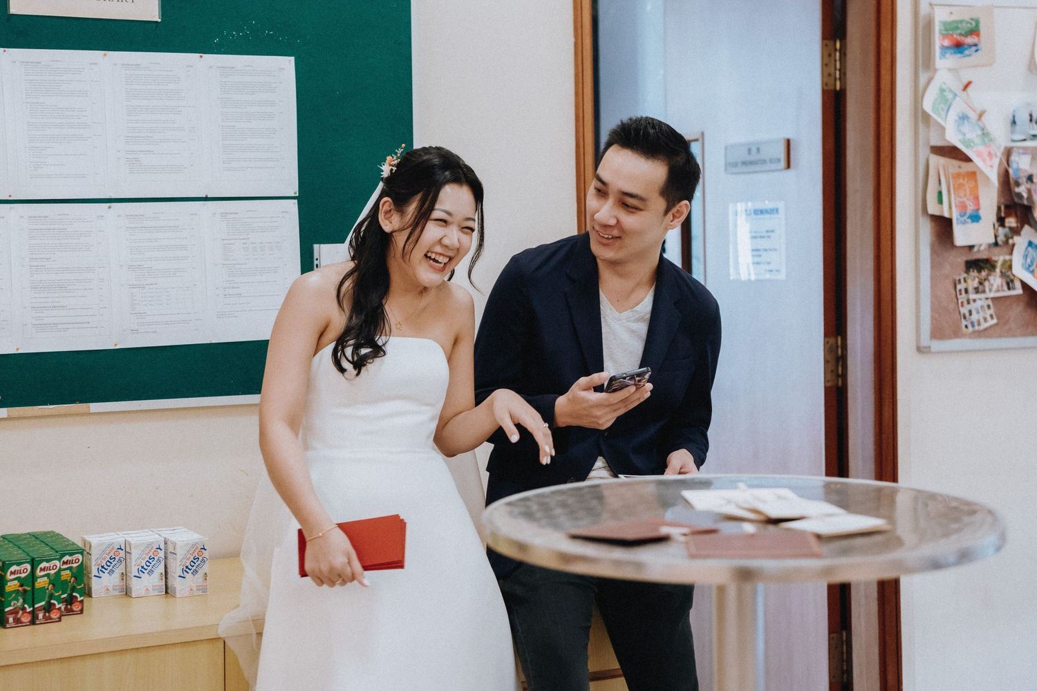 Bride in white dress and groom laugh together while looking at a phone at a table.