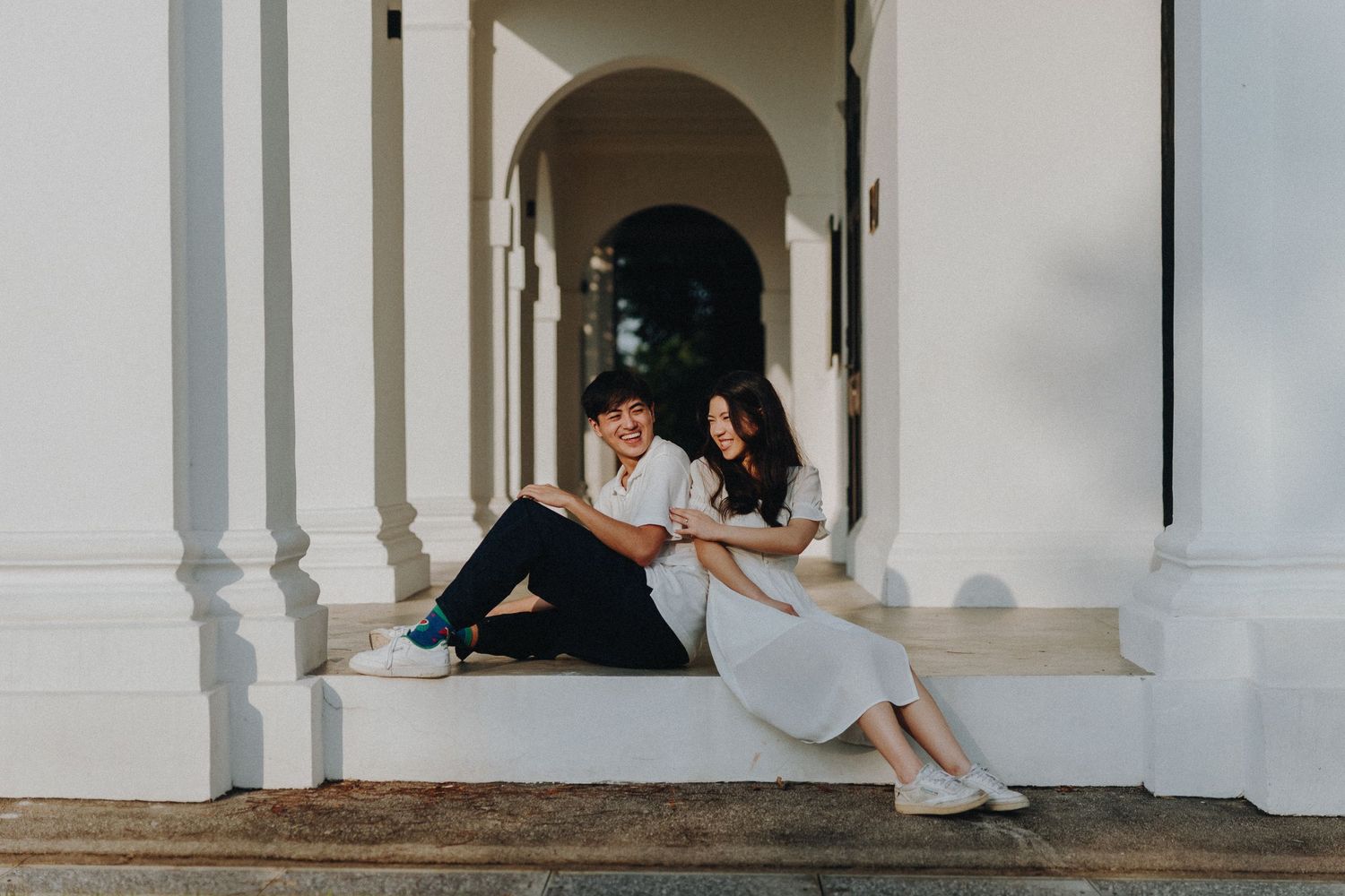 Couple in casual white outfits sitting together in an elegant white archway colonnade.
