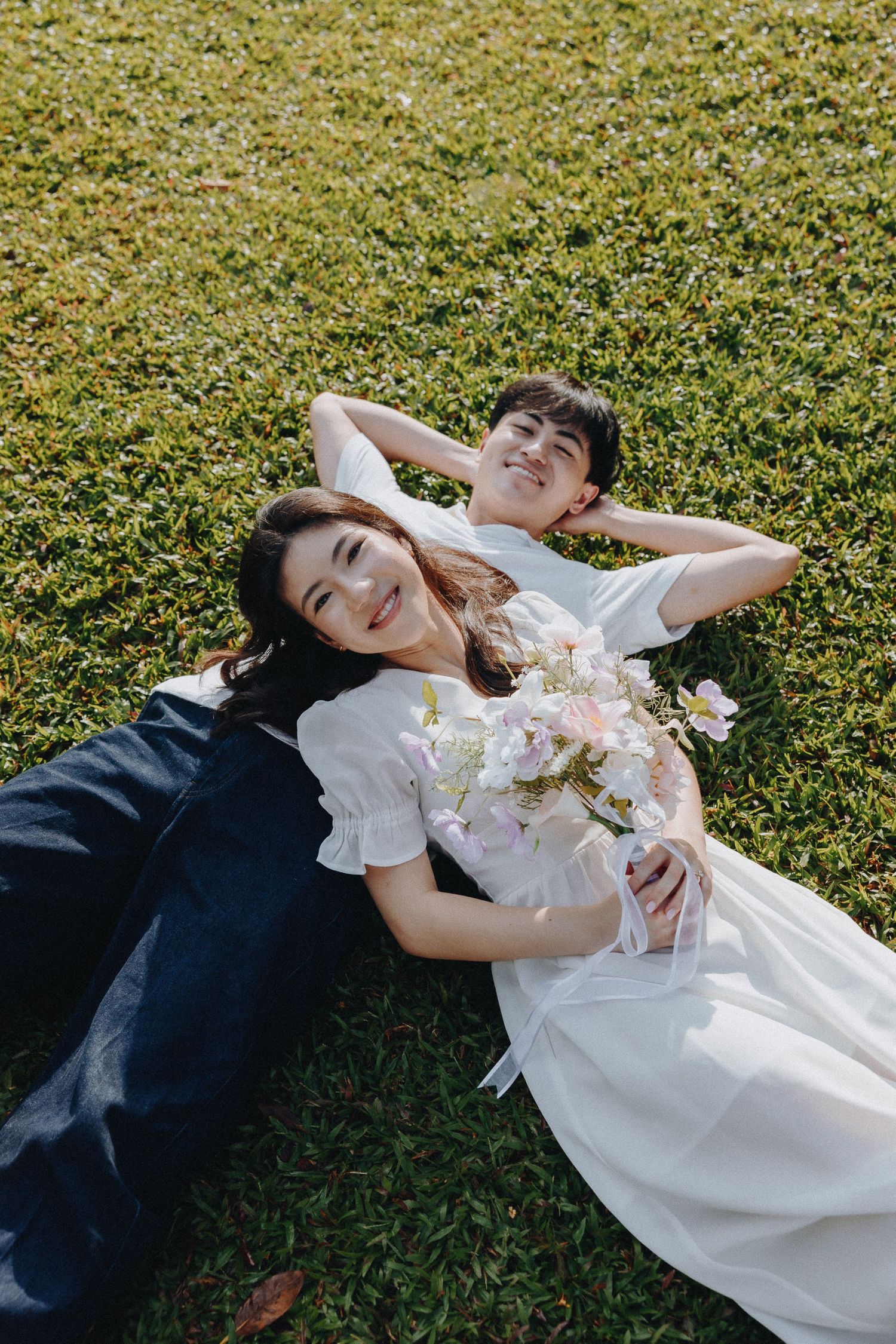 A couple laying on grass with flowers, sharing a tender moment outdoors.