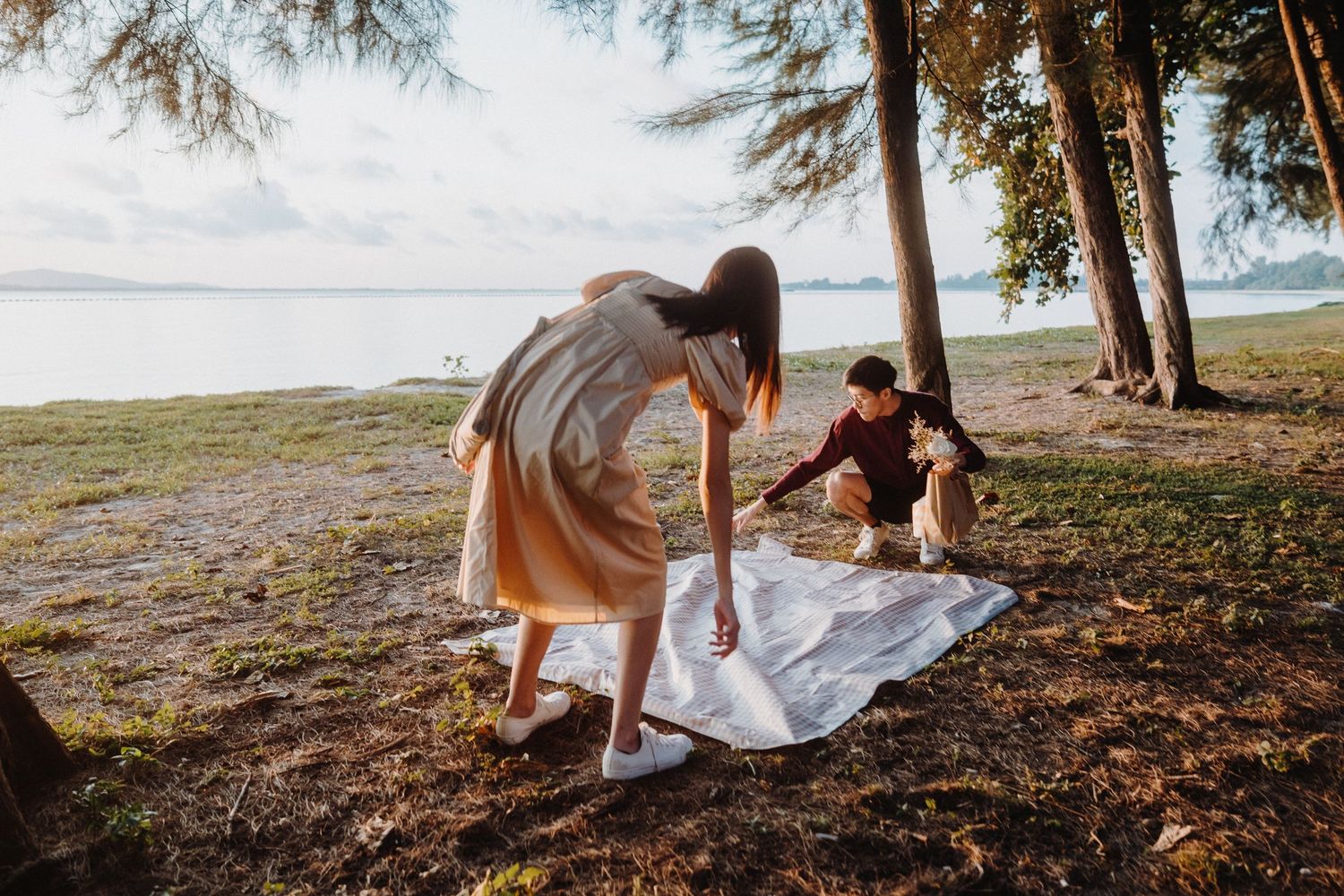 Couple enjoying a romantic picnic on a blanket under trees at sunset by the water.