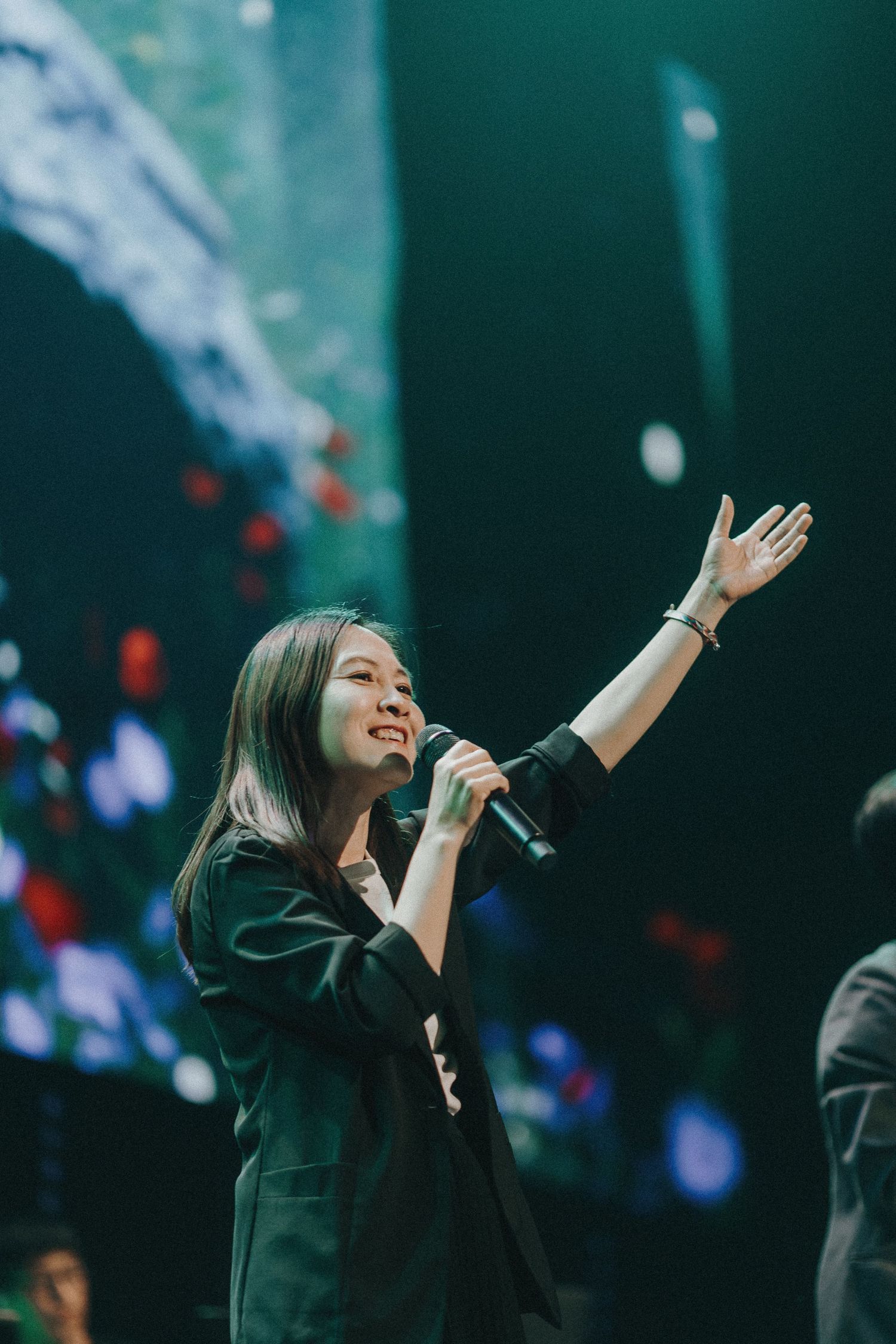 A performer in a black outfit raises their arms while singing passionately on stage with dramatic teal lighting.