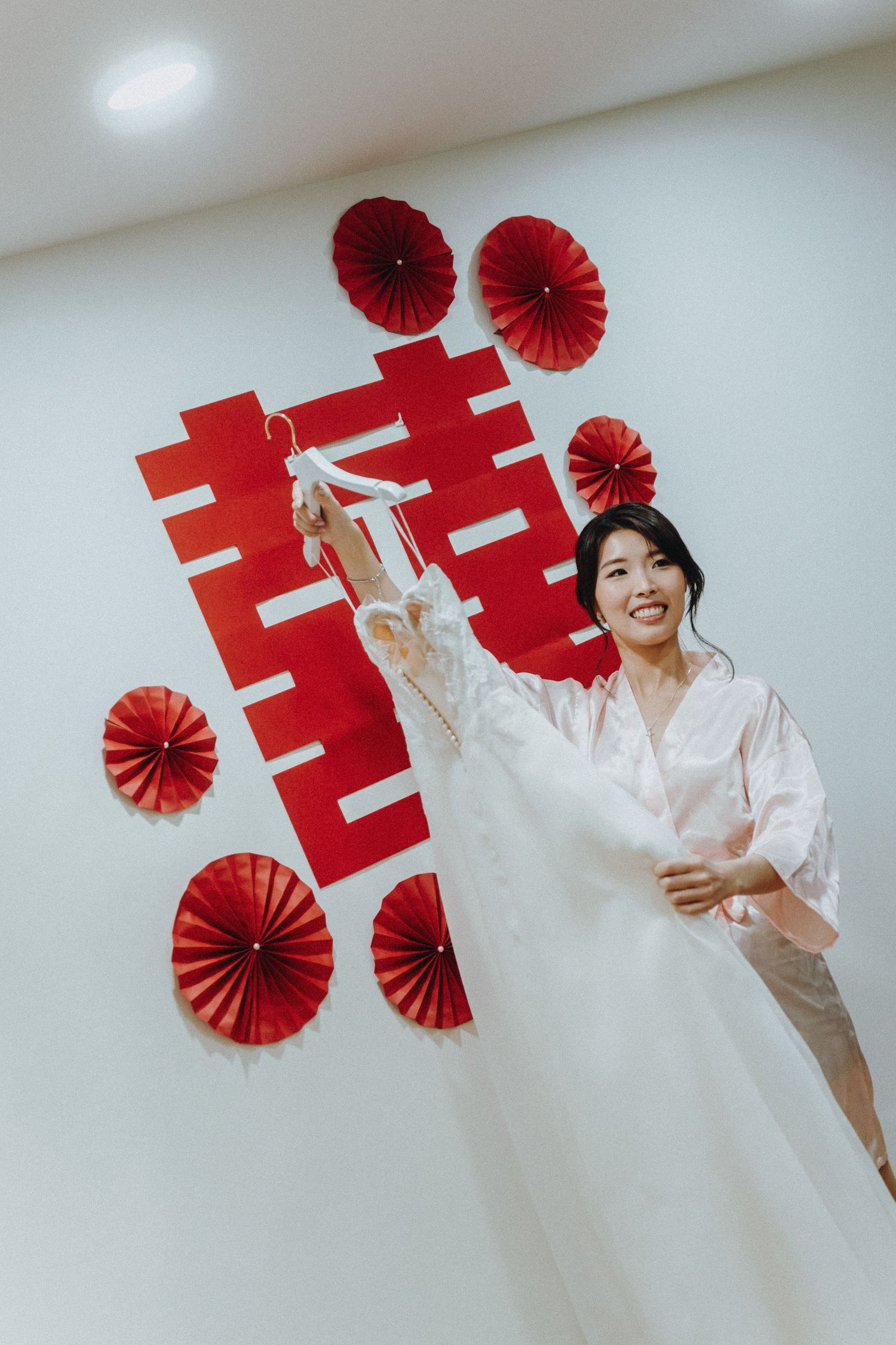Wedding preparation scene with traditional Chinese double happiness symbol and red paper fan wall decorations.