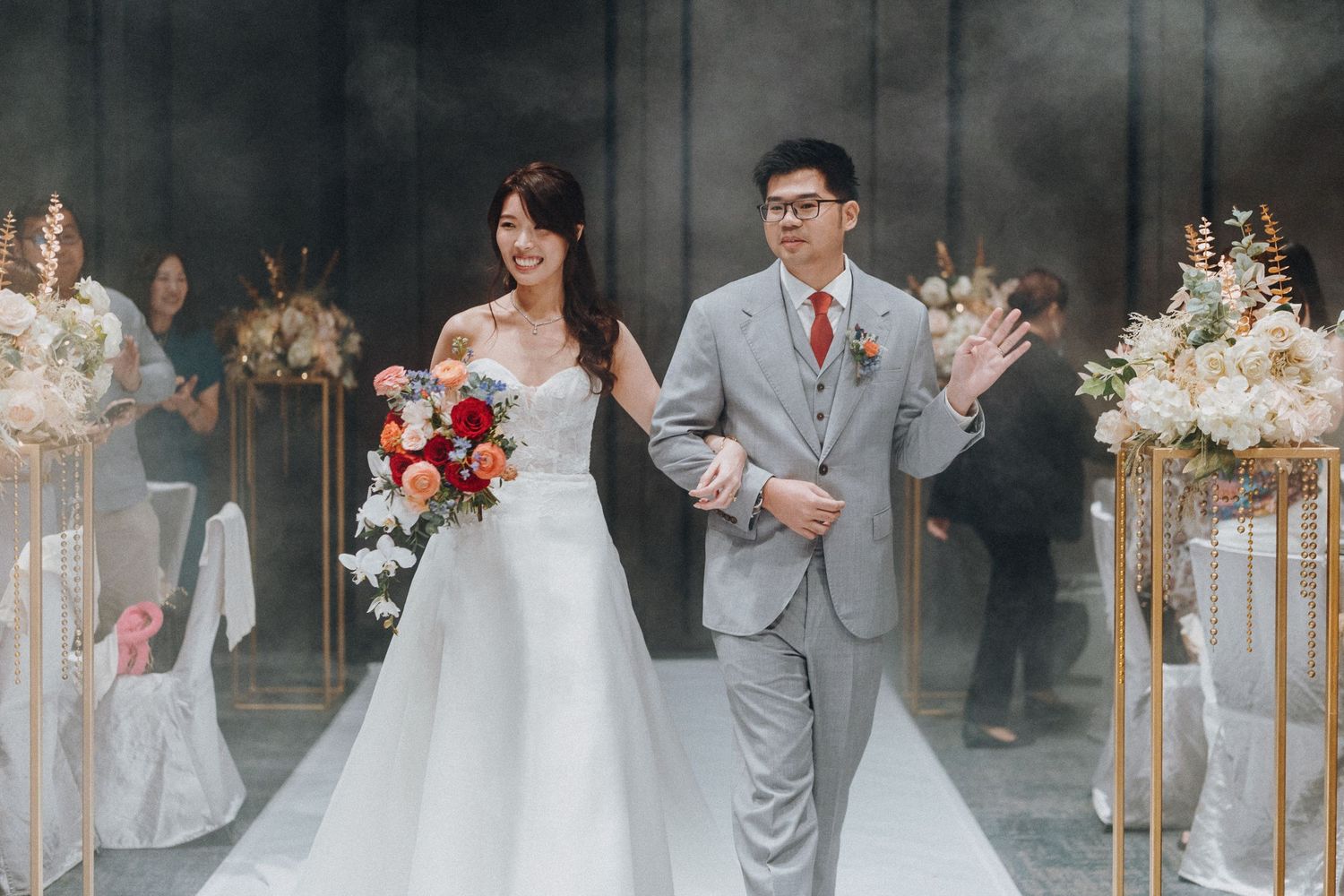 A couple walks down the aisle at their wedding ceremony with elegant floral decor and blurred guests in the background.