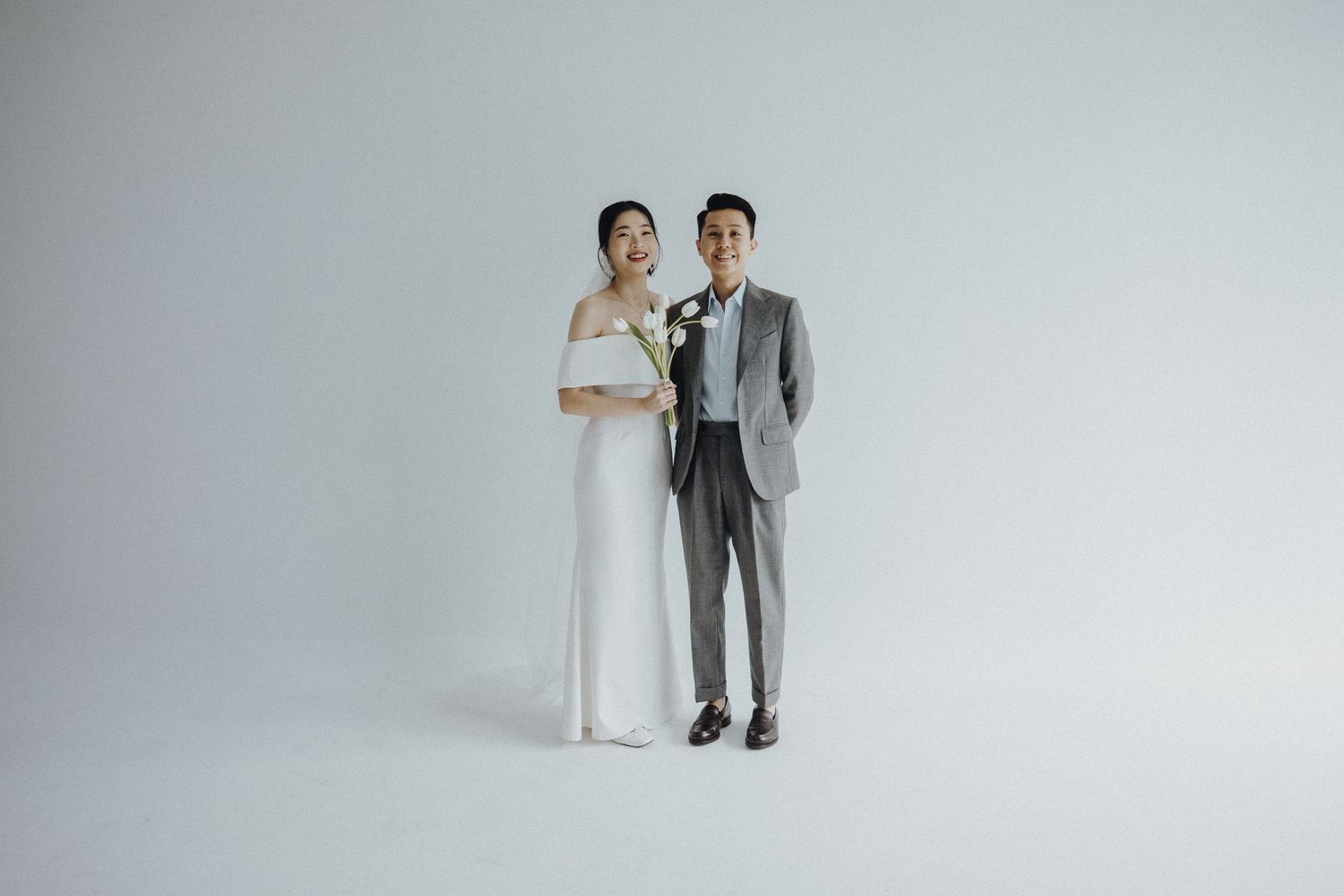 Couple in elegant wedding attire posing playfully against a minimal grey backdrop.