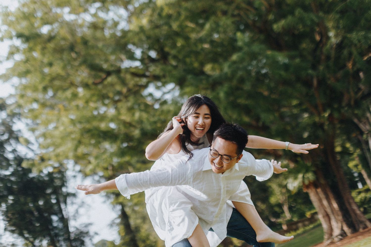 A couple in white clothing plays and laughs while giving a piggyback ride in a park.