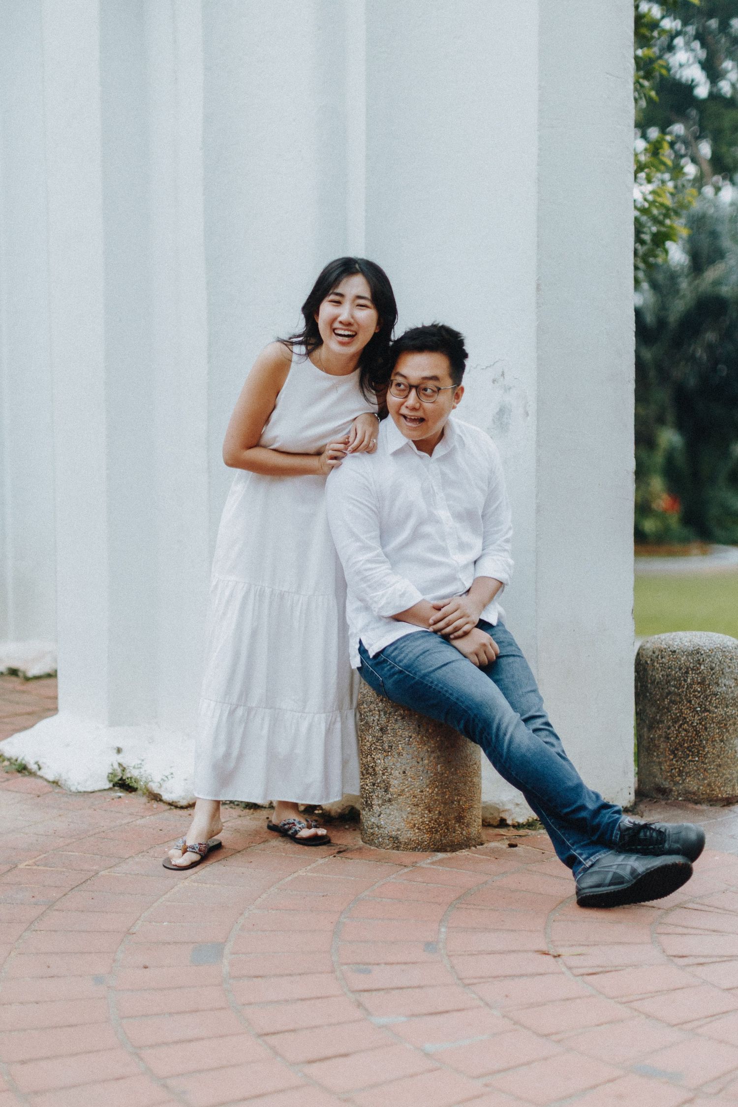 A couple in coordinated white outfits pose playfully against a classical white column on a brick path.