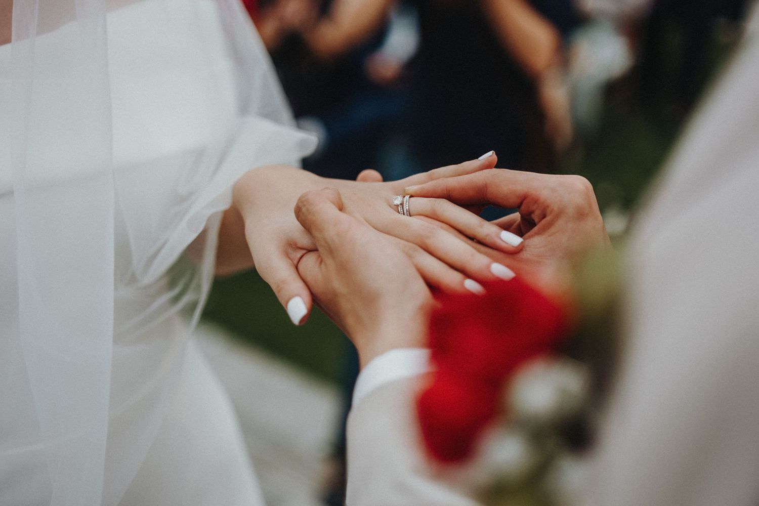 Close up view of hands exchanging a wedding ring during an outdoor ceremony with blurred guests in the background.