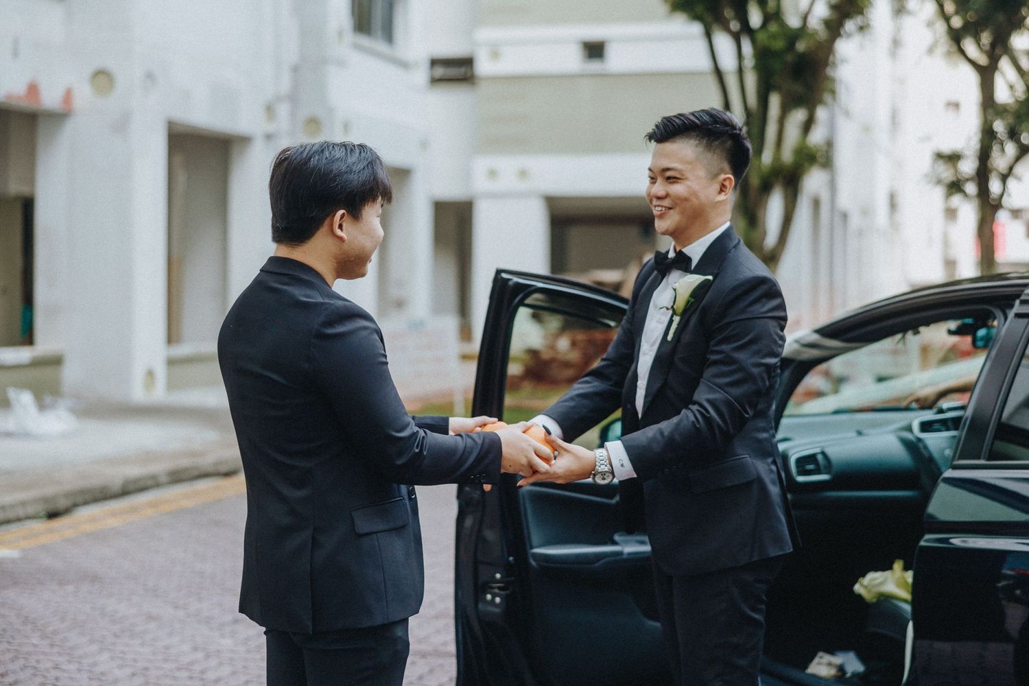Two businessmen in dark suits shake hands next to a black car on a quiet street.