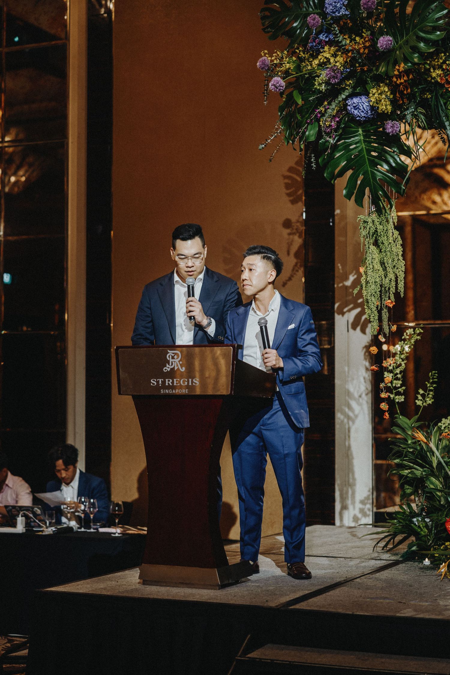 Two people in suits giving speeches at wedding reception podium with tropical floral arrangements.