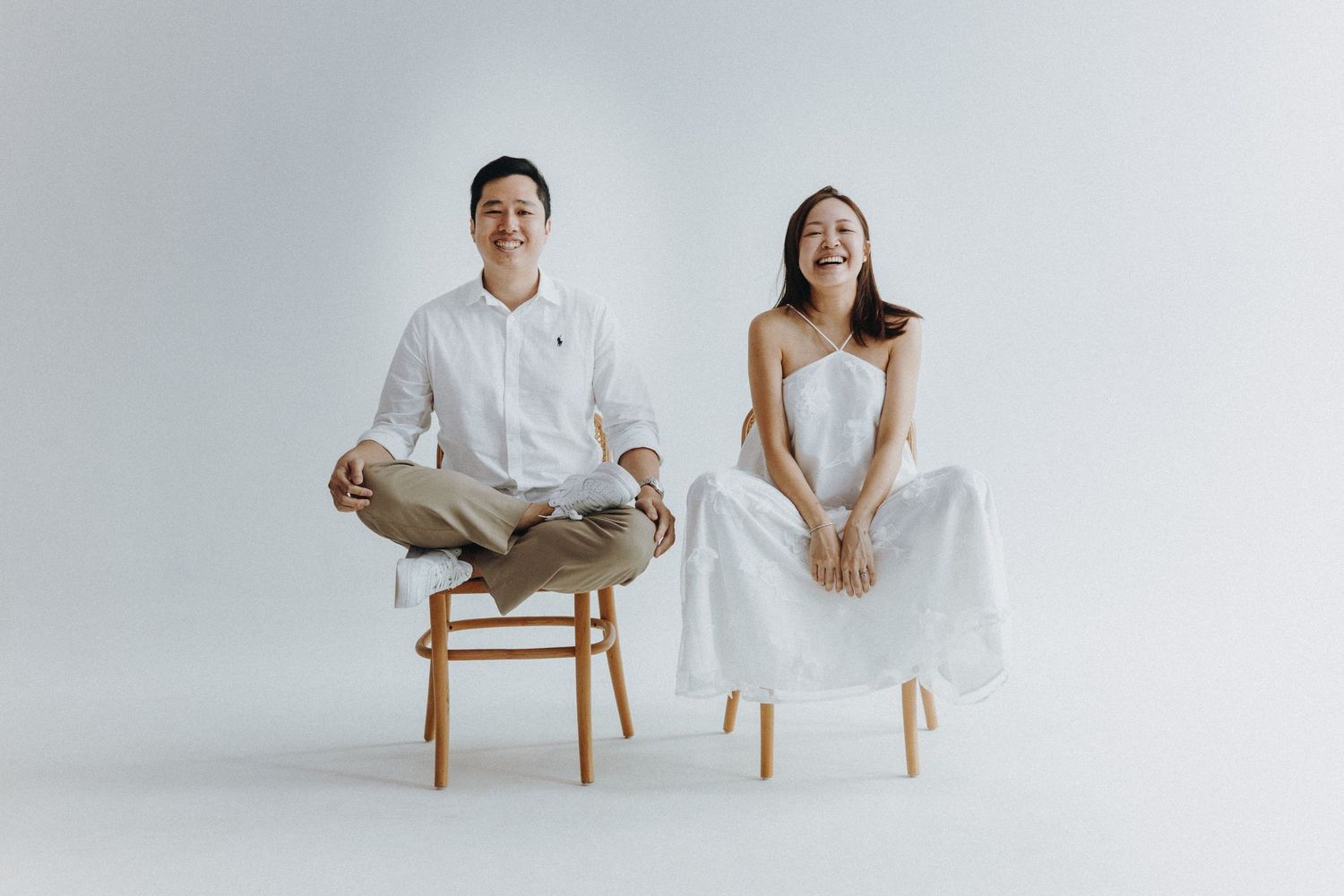 Minimalist studio portrait of couple in white attire sitting on wooden bench.