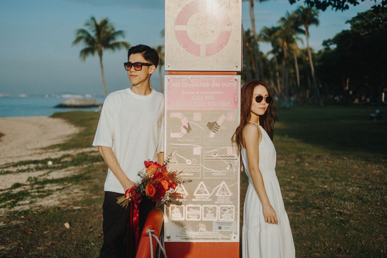Stylish couple in coordinated white outfits pose with red flowers near a beachside path with palm trees.
