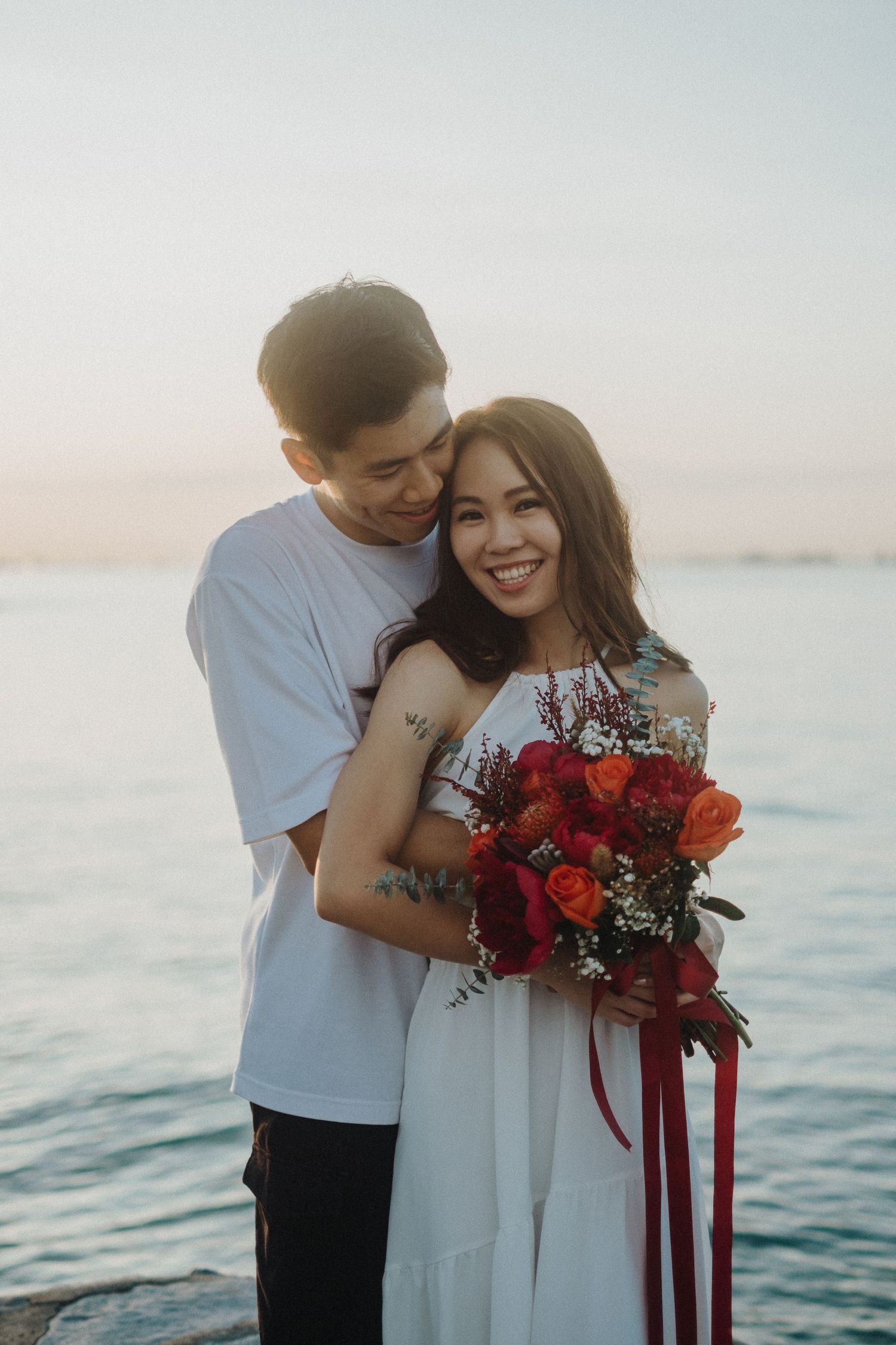 A romantic couple embraces by the ocean at sunset as she holds a vibrant red bouquet.