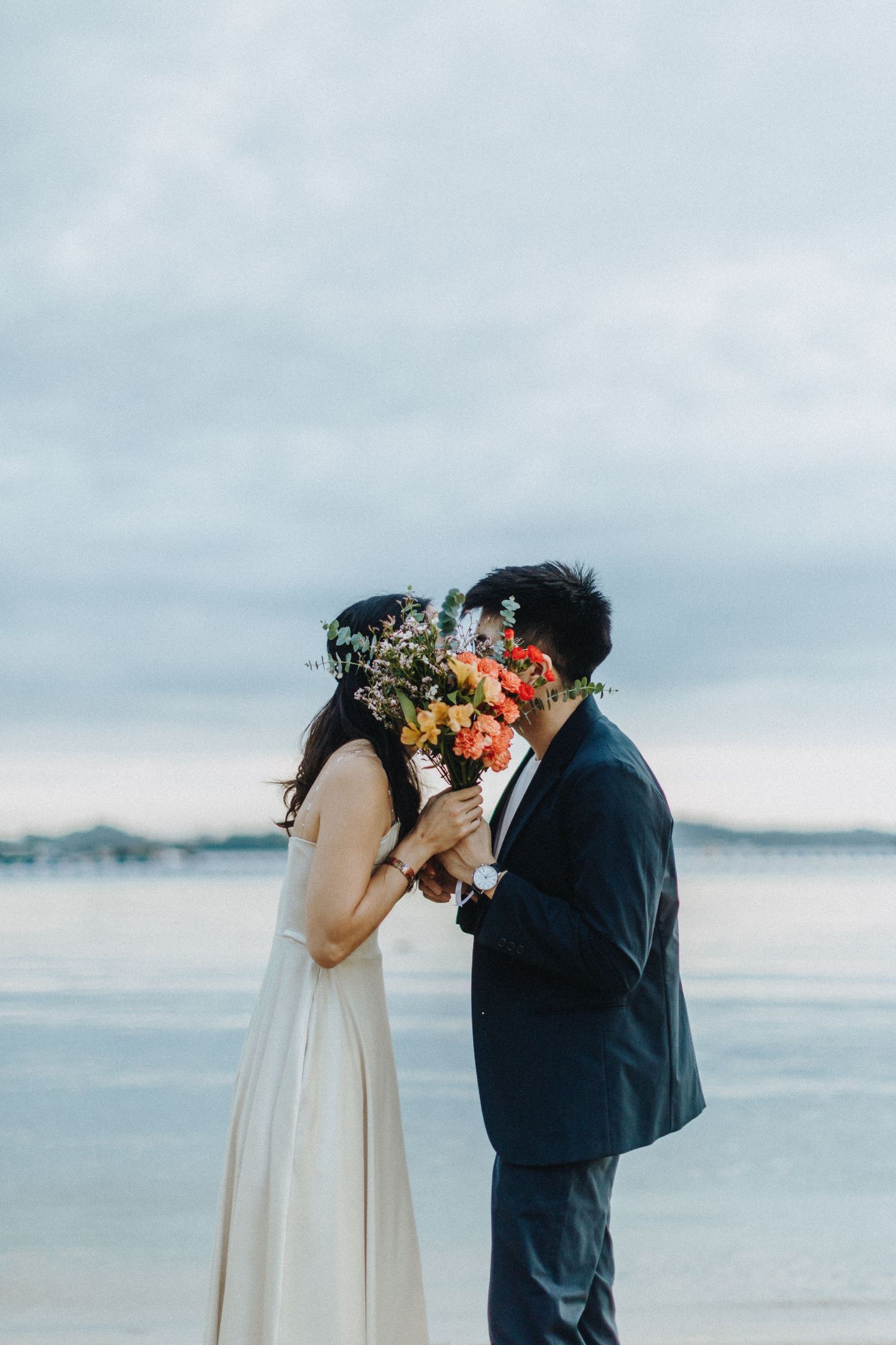 A romantic couple shares a moment behind a colorful bouquet against a moody coastal backdrop.