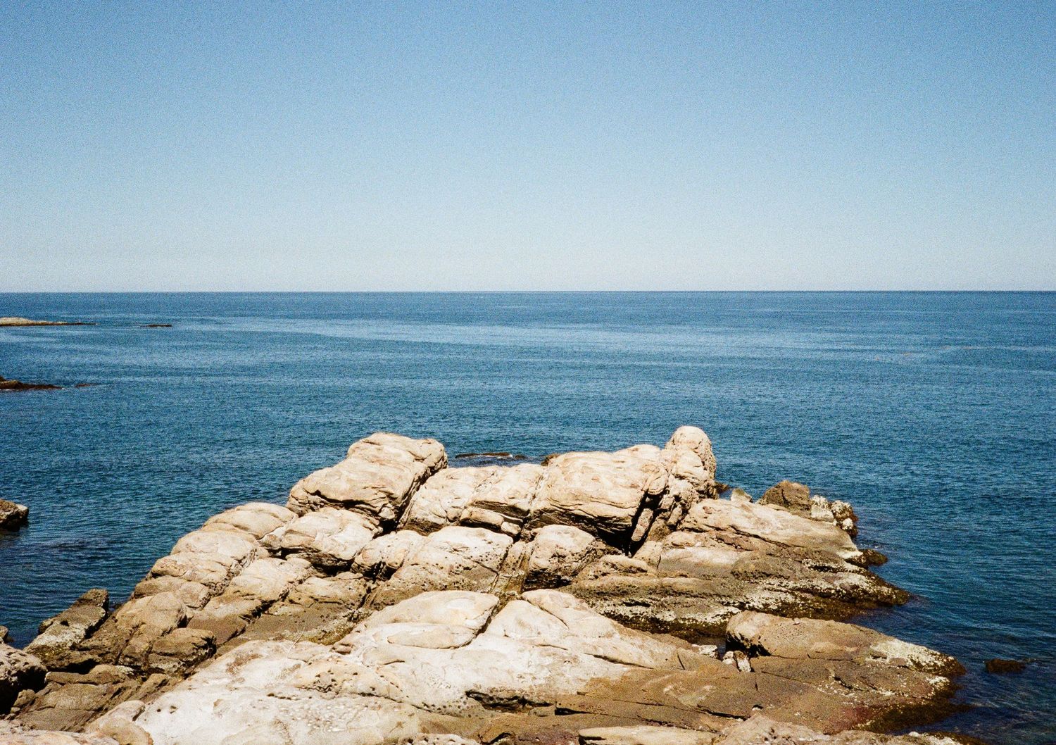 Large weathered rock formations jutting out into the calm blue ocean waters under clear skies.