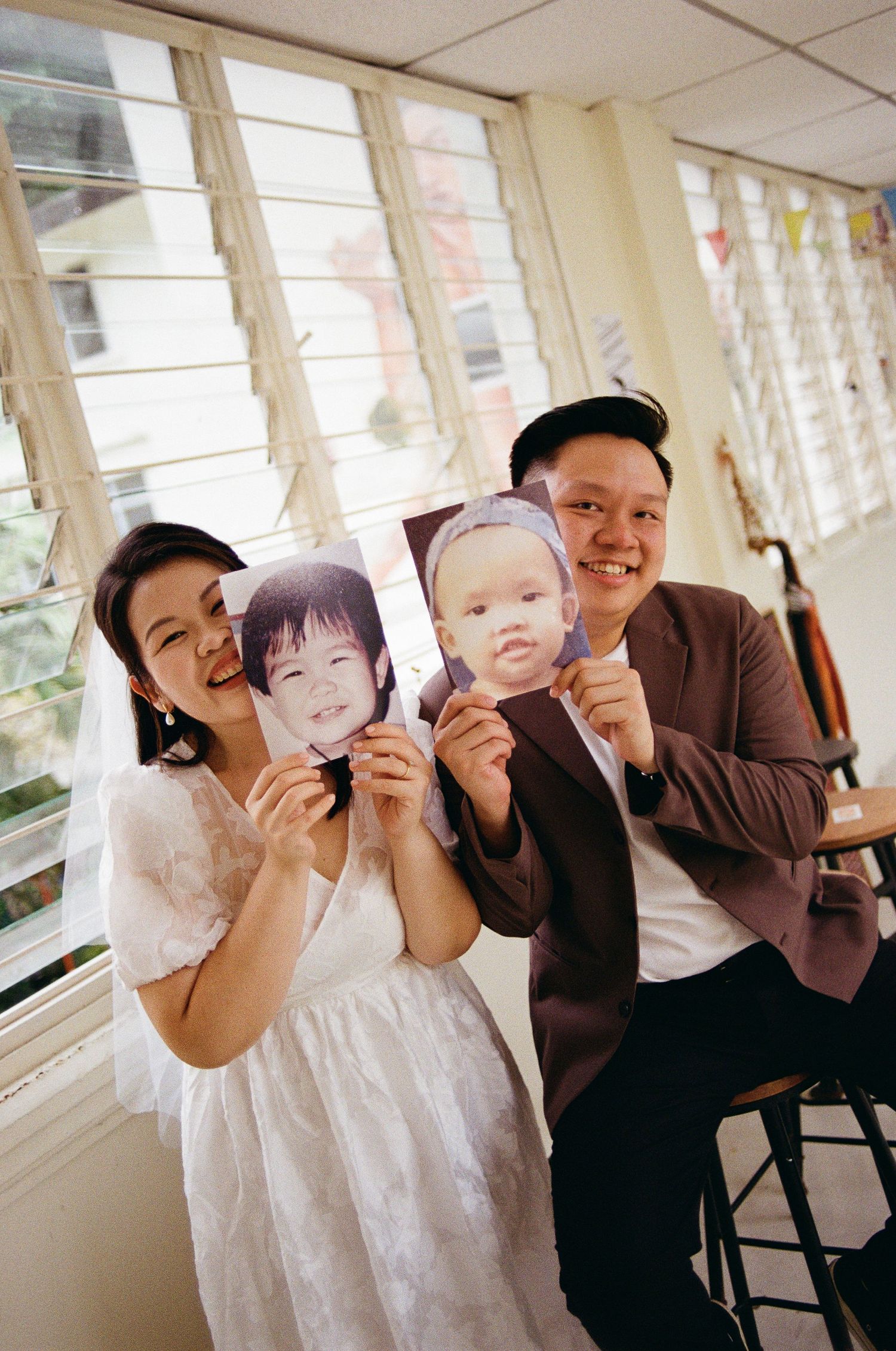 A couple holding up photographs while sitting in a bright hallway with louvered windows.