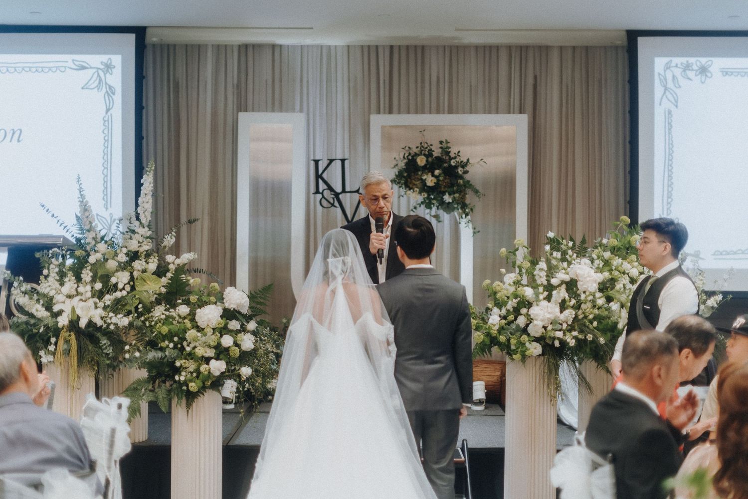 A wedding ceremony in progress with white floral arrangements and elegant drapes decorating the venue.