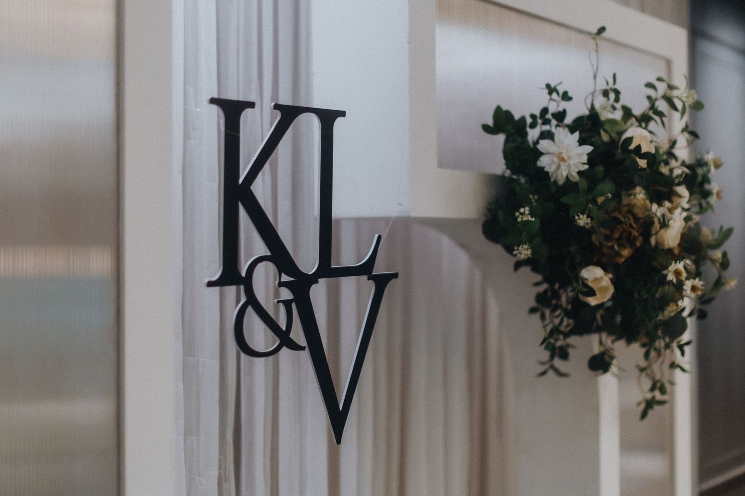 Black monogram letters KL&V displayed on a white wall with decorative greenery and flowers.
