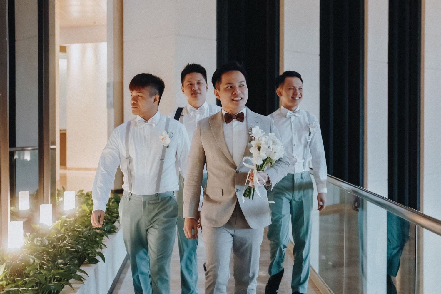 A group of formally dressed men in light colored attire walk together along a sunlit hallway for a wedding celebration.
