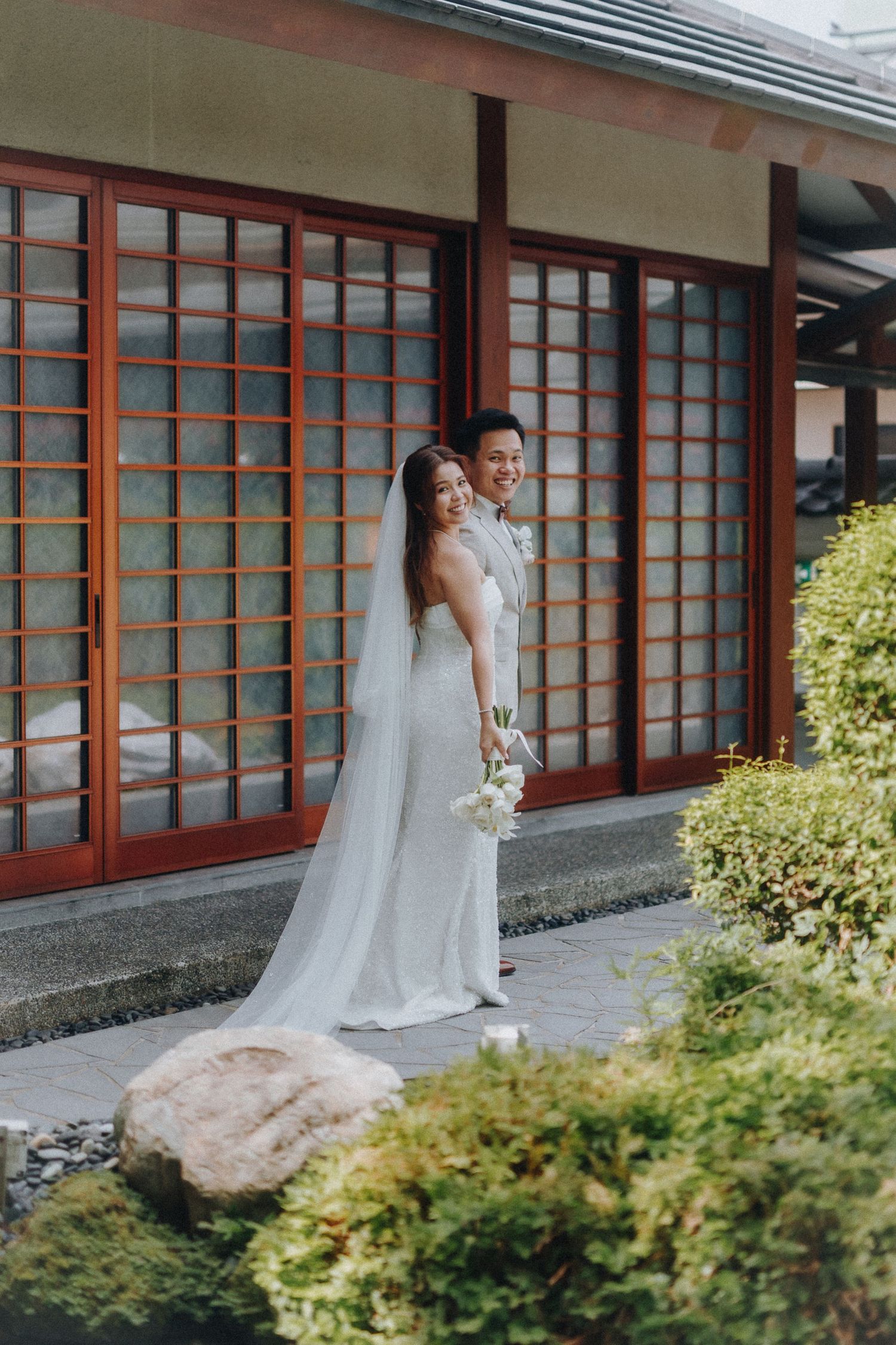 Elegant couple in wedding attire poses by Japanese-style building with garden landscaping.