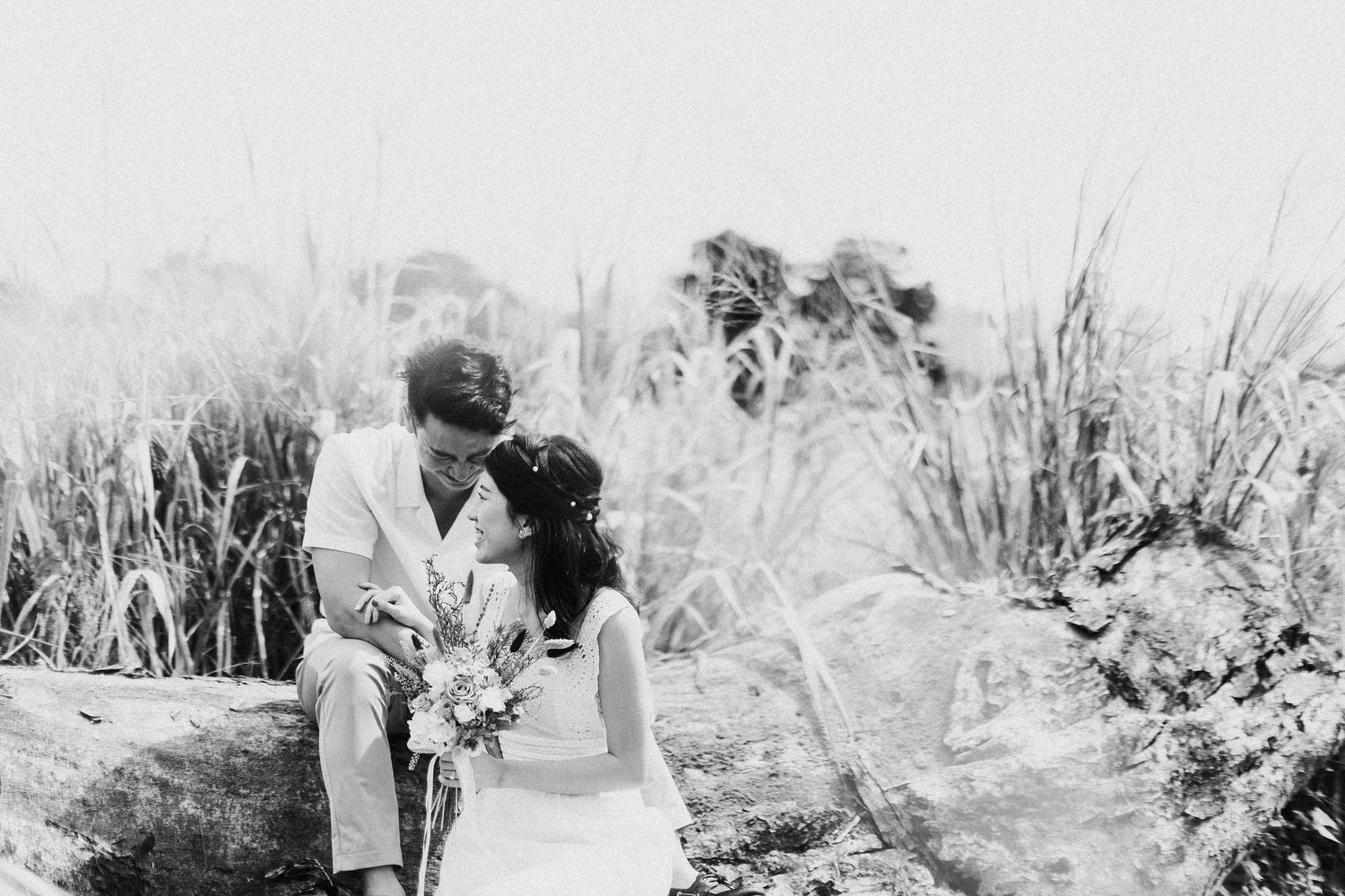 A romantic black and white photo of a couple embracing in a field with tall grass and natural scenery.