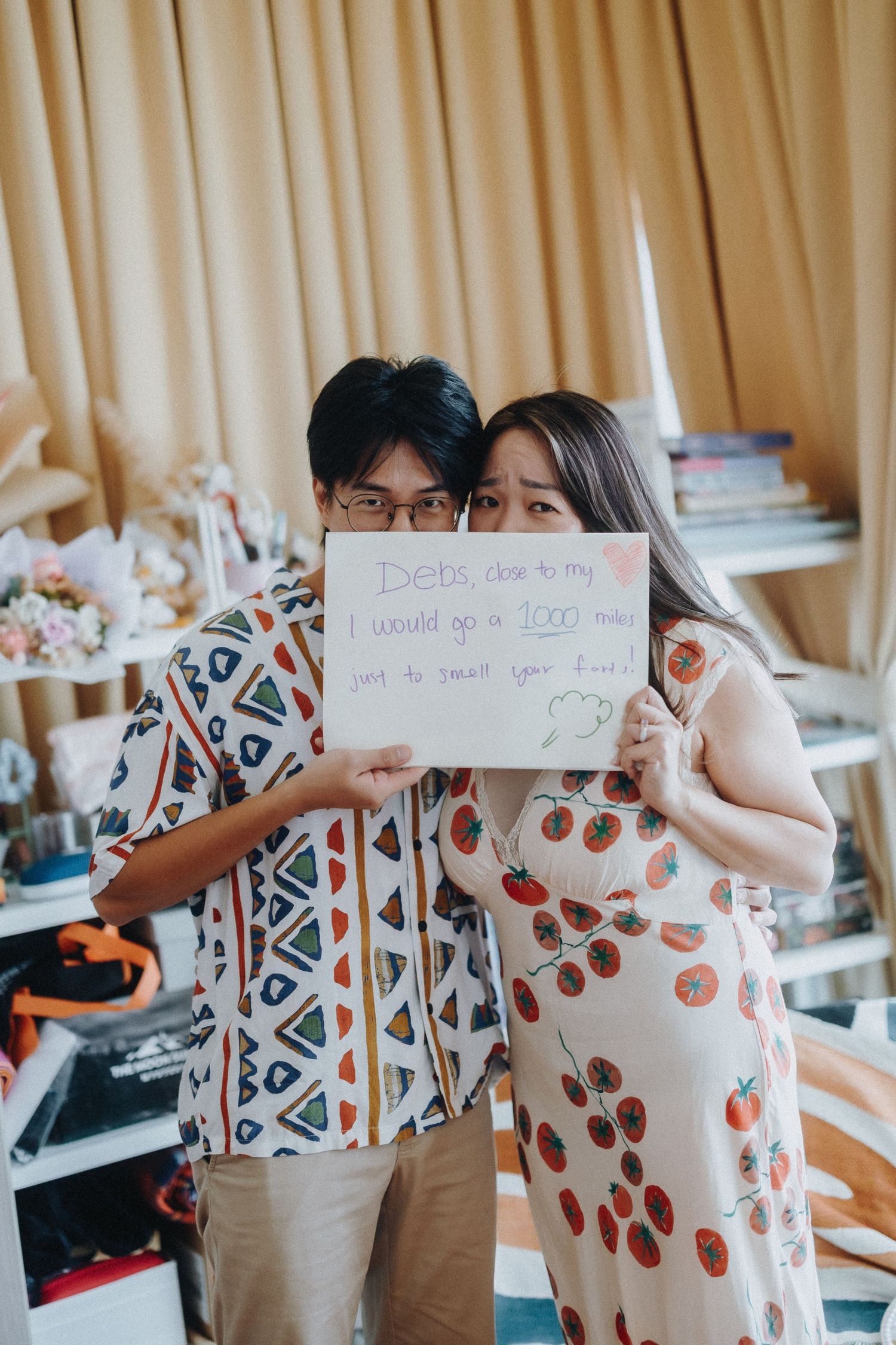 A couple holds a handwritten note while posing together in a room with beige curtains and decorative elements.