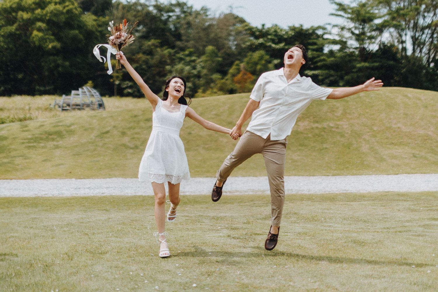 Two people in matching white outfits jumping and playing together on a sunny grassy hill.