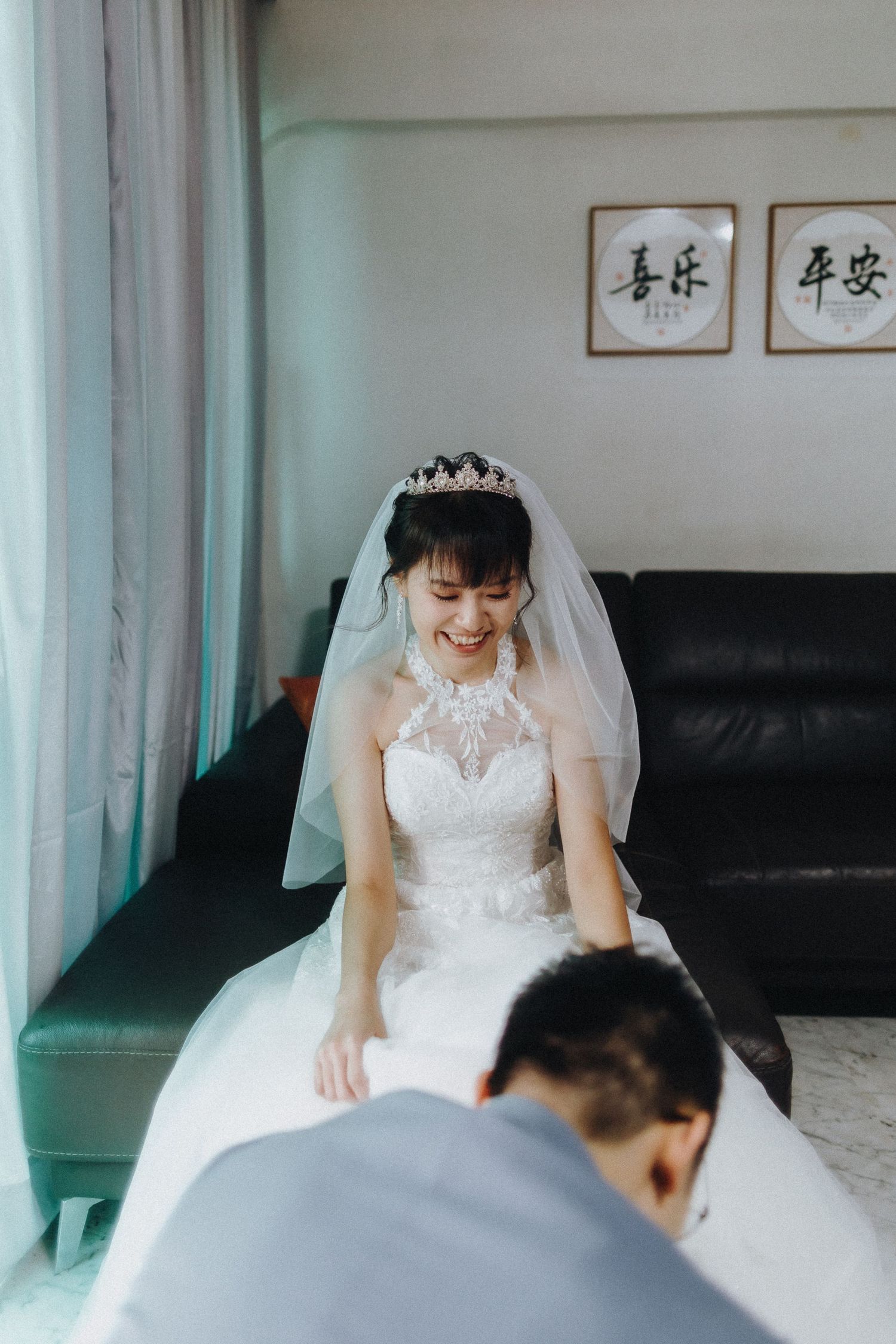 A bride in a white lace wedding dress and veil sits on a green sofa beneath Chinese calligraphy wall art.