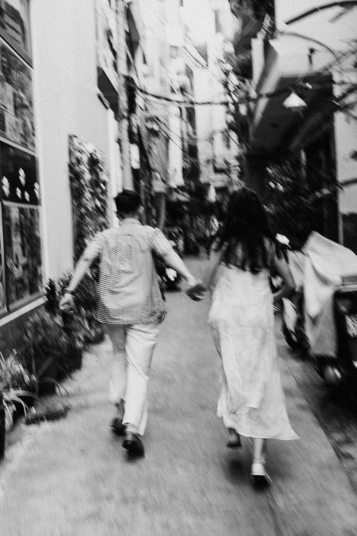 A black and white motion blur photograph shows two people walking down a narrow urban street.