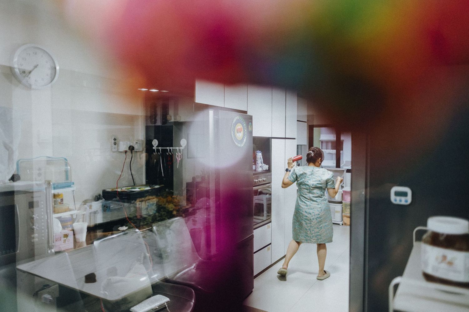 A colorful blurred foreground frames a view into a modern white kitchen where someone is working.
