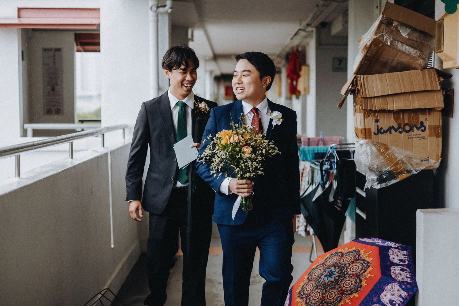 Two people in formal suits share a joyful moment in a corridor while one holds a bouquet of yellow flowers.