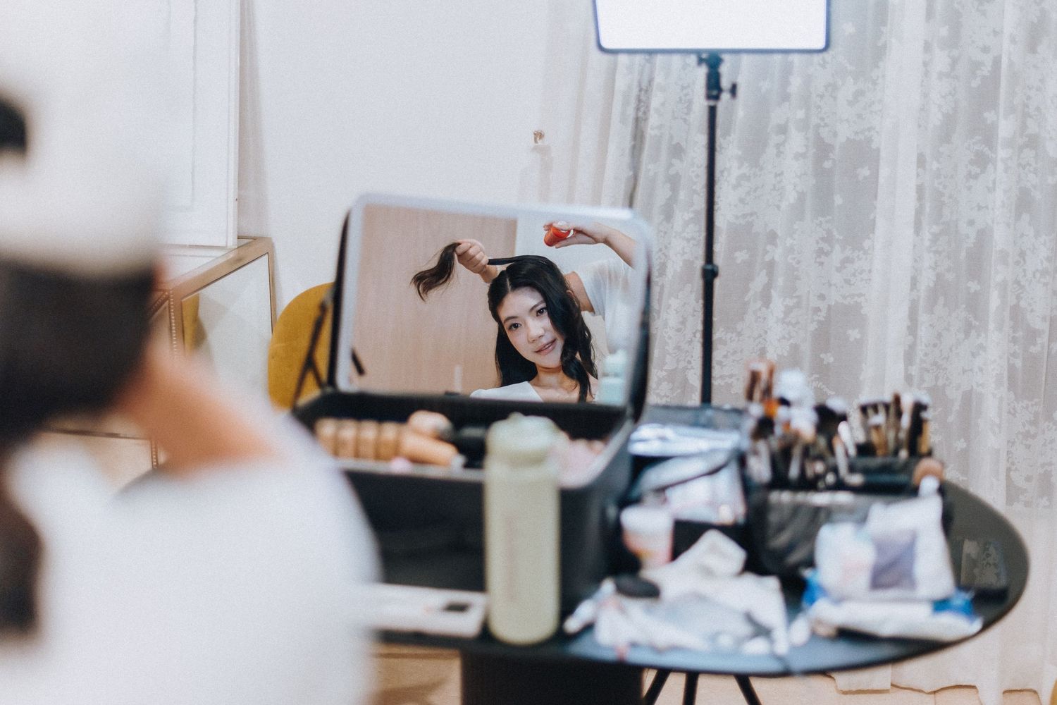 Woman applying makeup while looking into a lighted vanity mirror surrounded by beauty products on a black table.