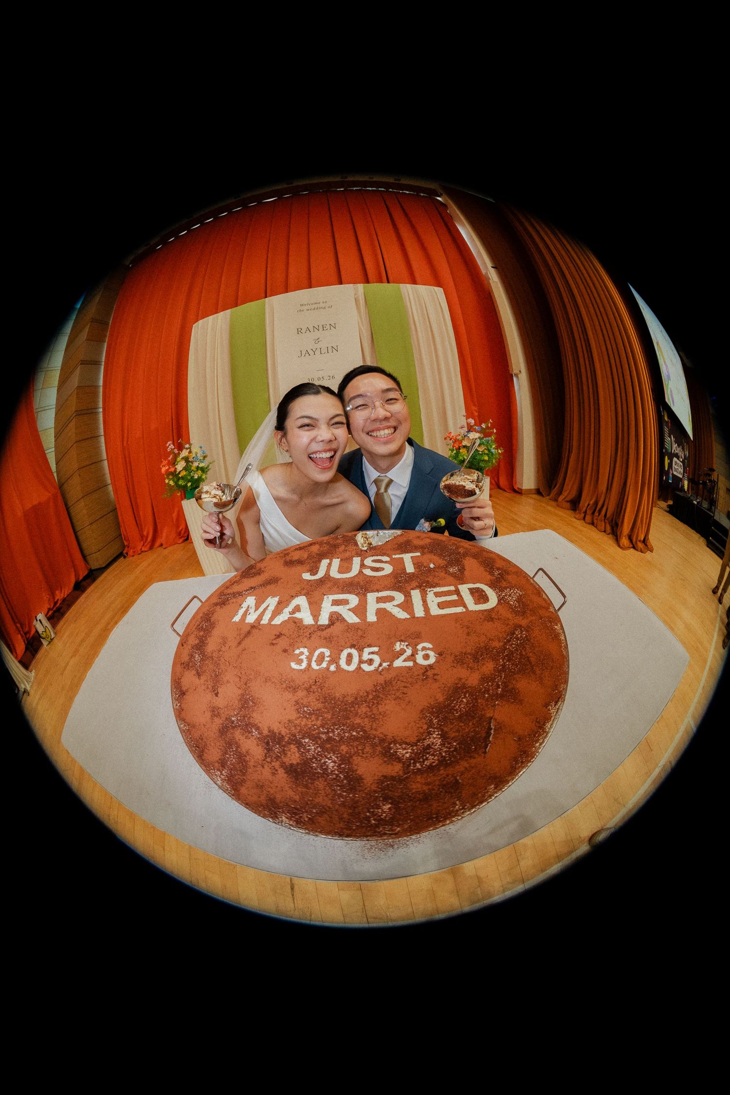 Newlyweds pose behind a Just Married cake with date 30.05.26 in a fisheye lens wedding photo.