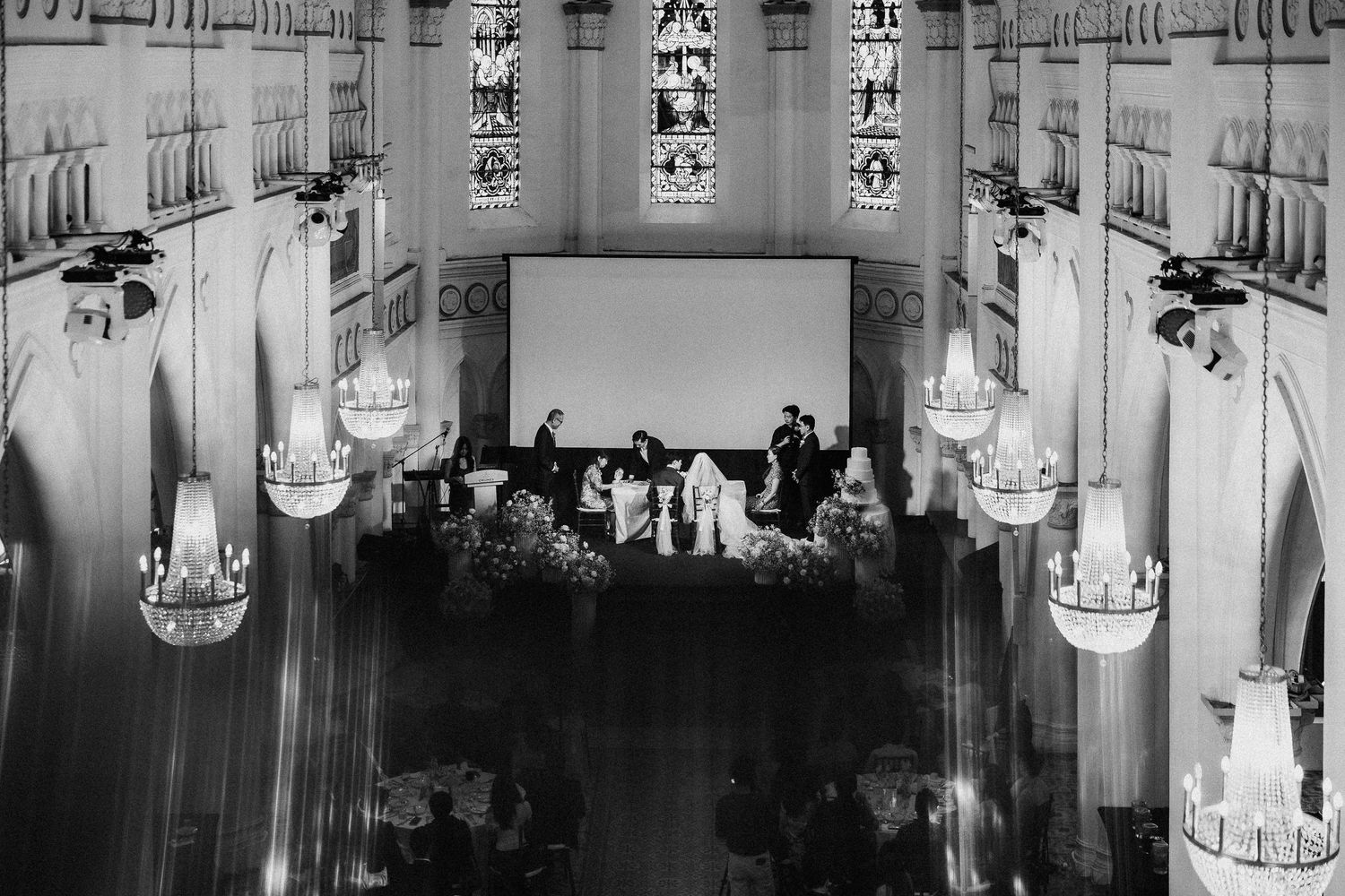 Black and white photo of an elegant church wedding ceremony with chandeliers and stained glass windows.
