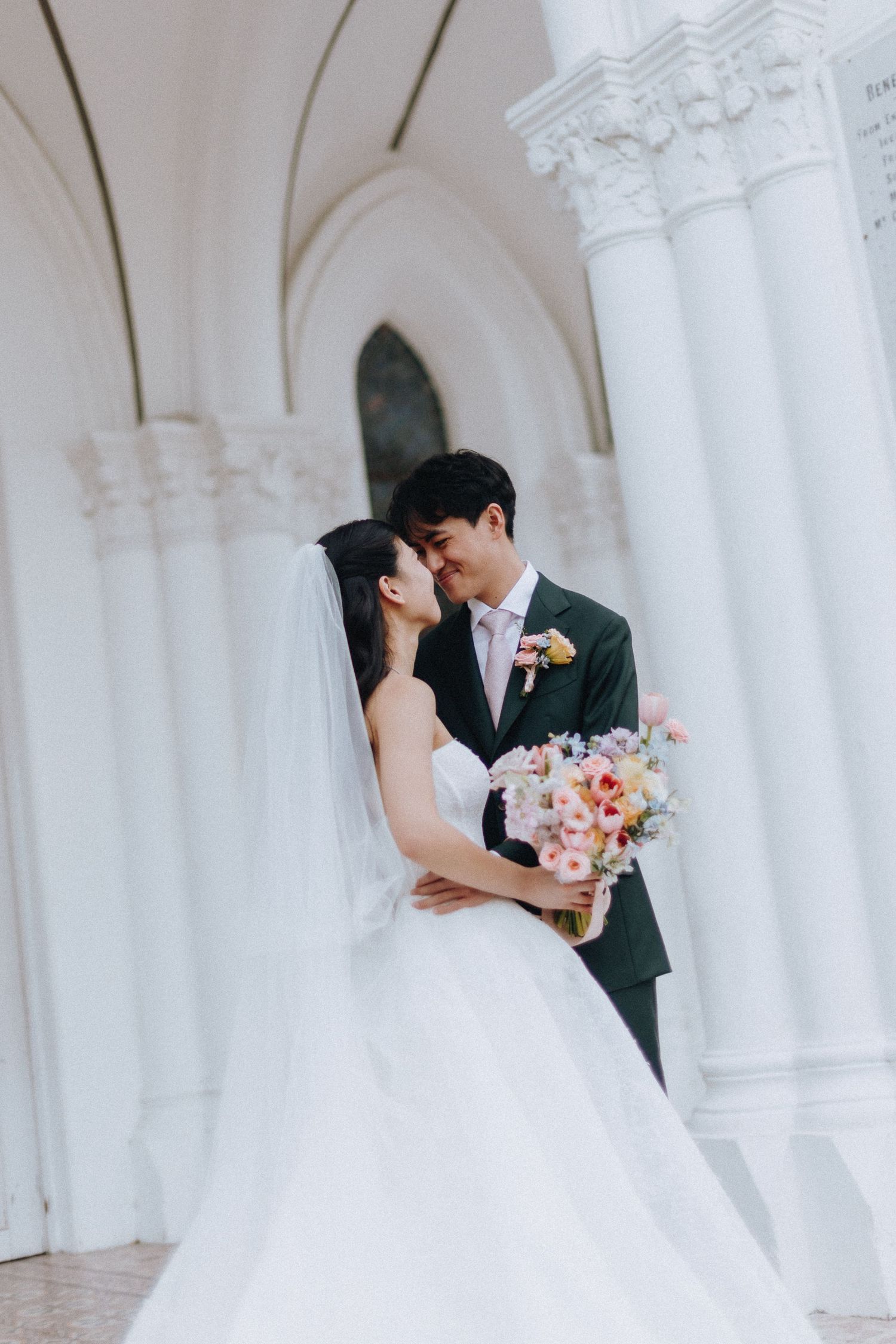 A bride and groom share a romantic moment in front of elegant white columns, the bride holding a colorful floral bouquet.