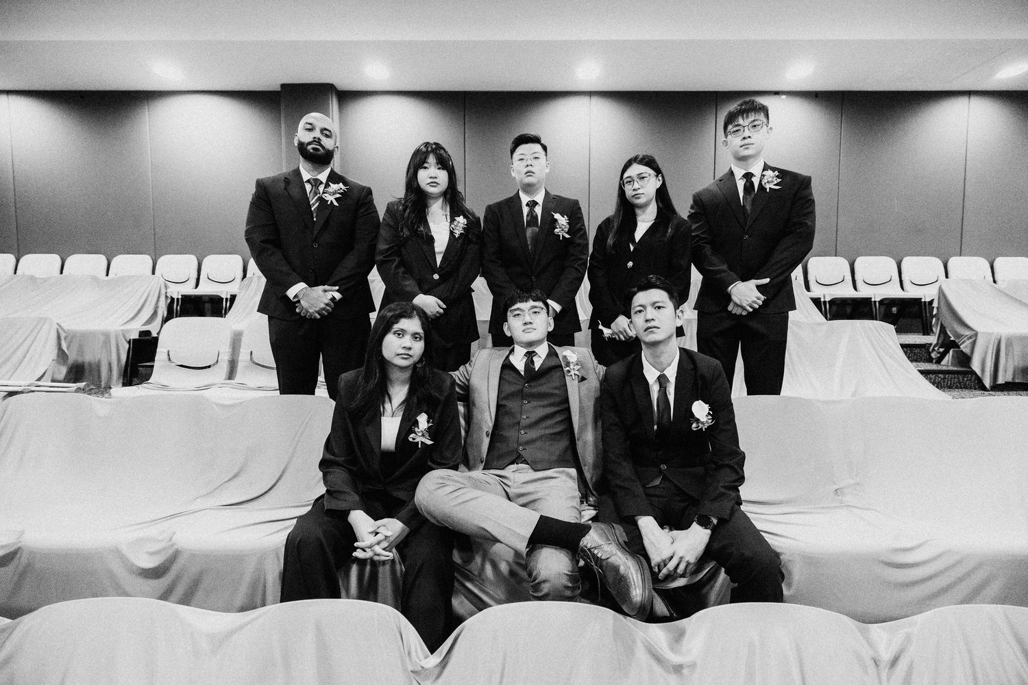 A black and white portrait of a wedding party posed in rows of white chairs.