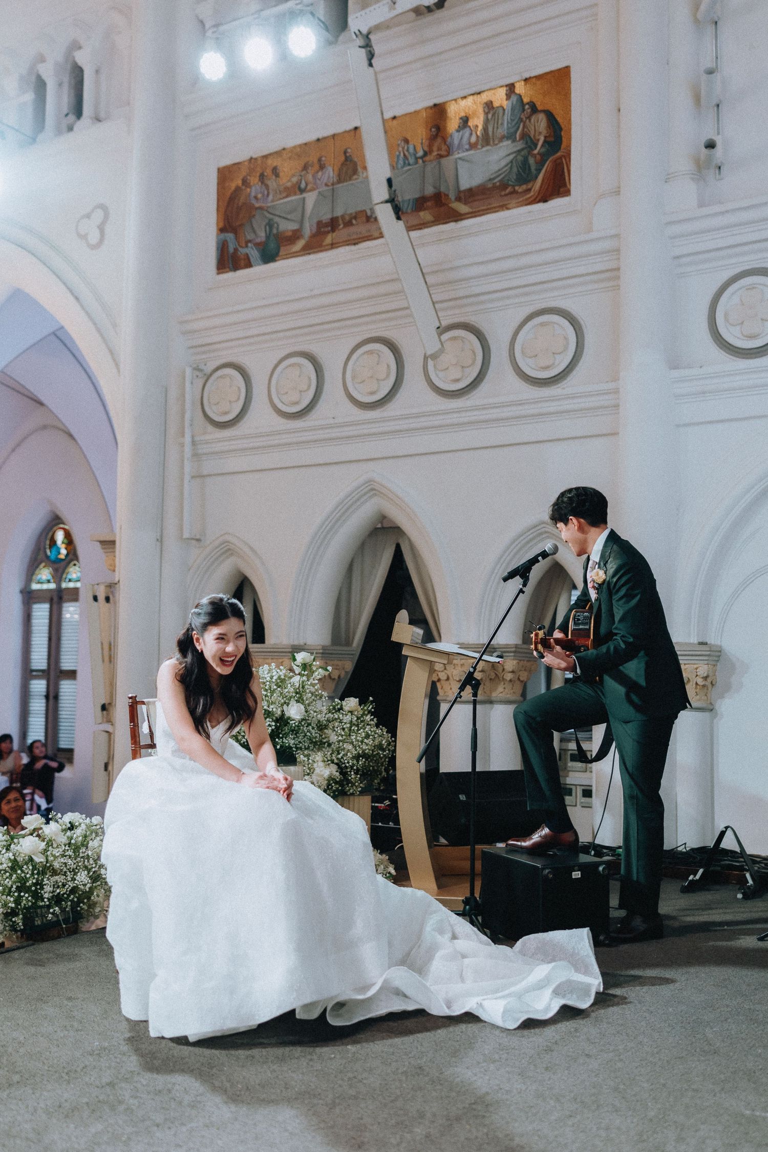 Bride in white gown sits listening as groom plays acoustic guitar and sings at their church wedding ceremony.