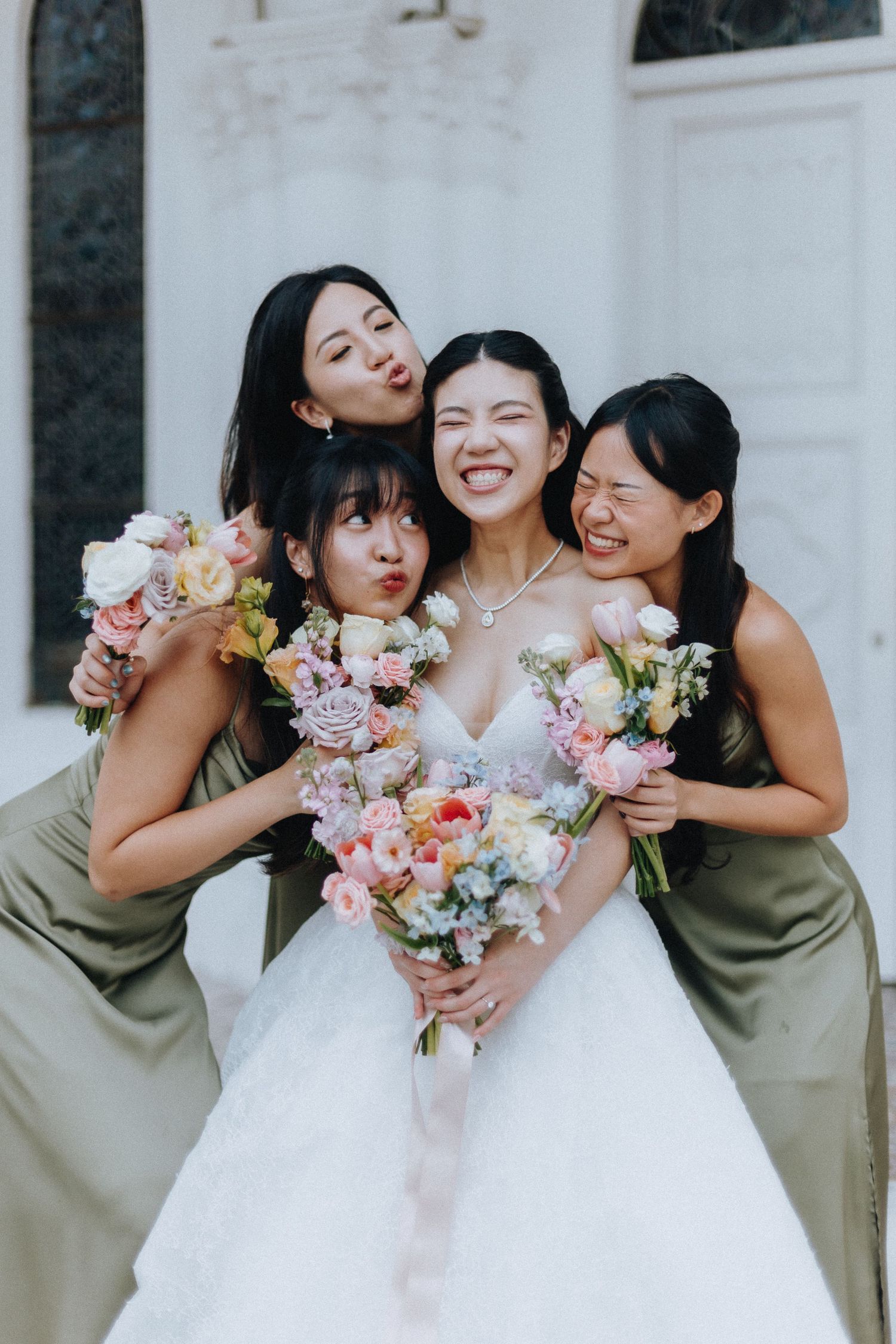 Bride in white gown holds floral bouquet with three bridesmaids in olive dresses, all smiling and posing playfully.